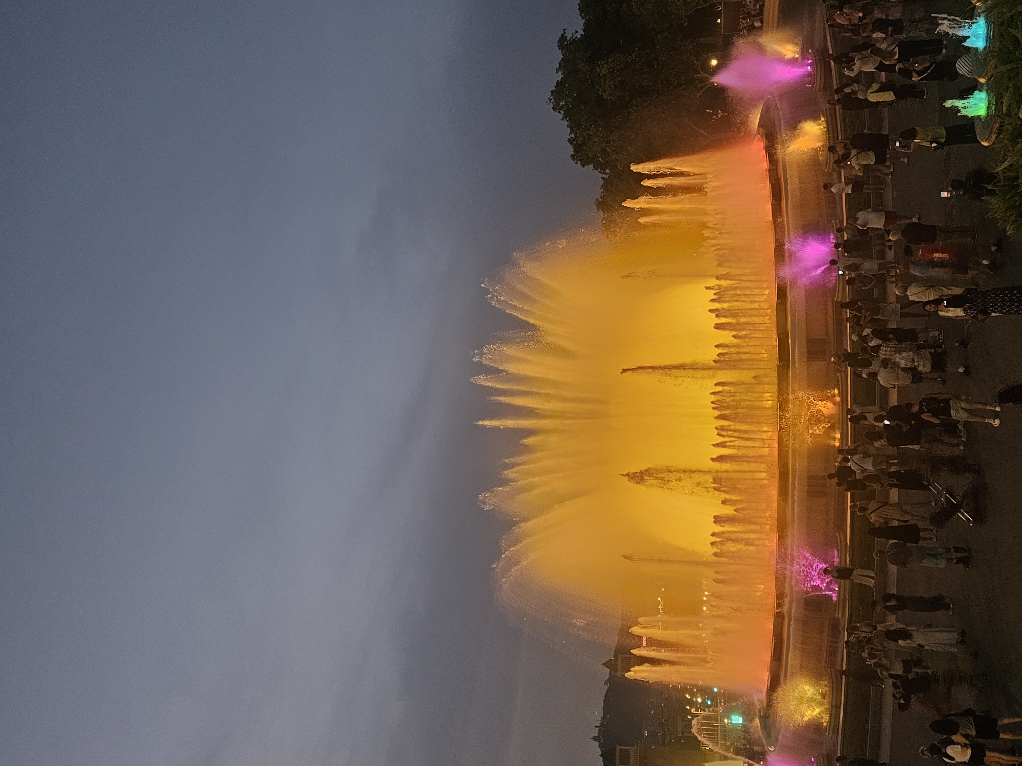 The image captures the Magic Fountain of Montjuïc in Barcelona, Spain, at dusk. A large, multi-tiered fountain dominates the scene, illuminated by vibrant yellow, orange, and pink lights that reflect off the water. Tall plumes of water shoot upwards, creating a dynamic display. In the foreground, a crowd of people stands gathered, observing the fountain. They are casually dressed, and some are holding up their phones to take pictures or videos. The sky above is a dusky blue, with some clouds visible. Trees line the background on the right side of the fountain, and in the distance, on the left, the silhouette of buildings and hills can be seen against the fading light. The overall atmosphere is one of awe and enjoyment of a public spectacle.