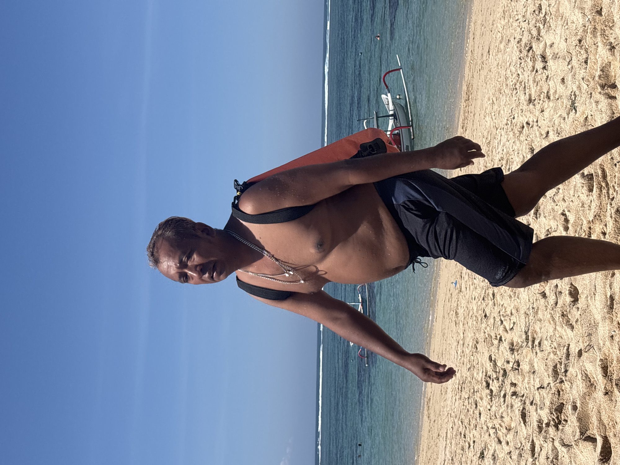 A man with graying hair, bare-chested, wearing dark shorts and a backpack with an orange bag, walks on a sandy beach in Kuta Selatan, Indonesia. The ocean, with calm blue water and small white waves, is visible in the background. Several small boats are anchored offshore, and a few people can be seen in the water. The sky is clear and blue. The man is looking to his left.