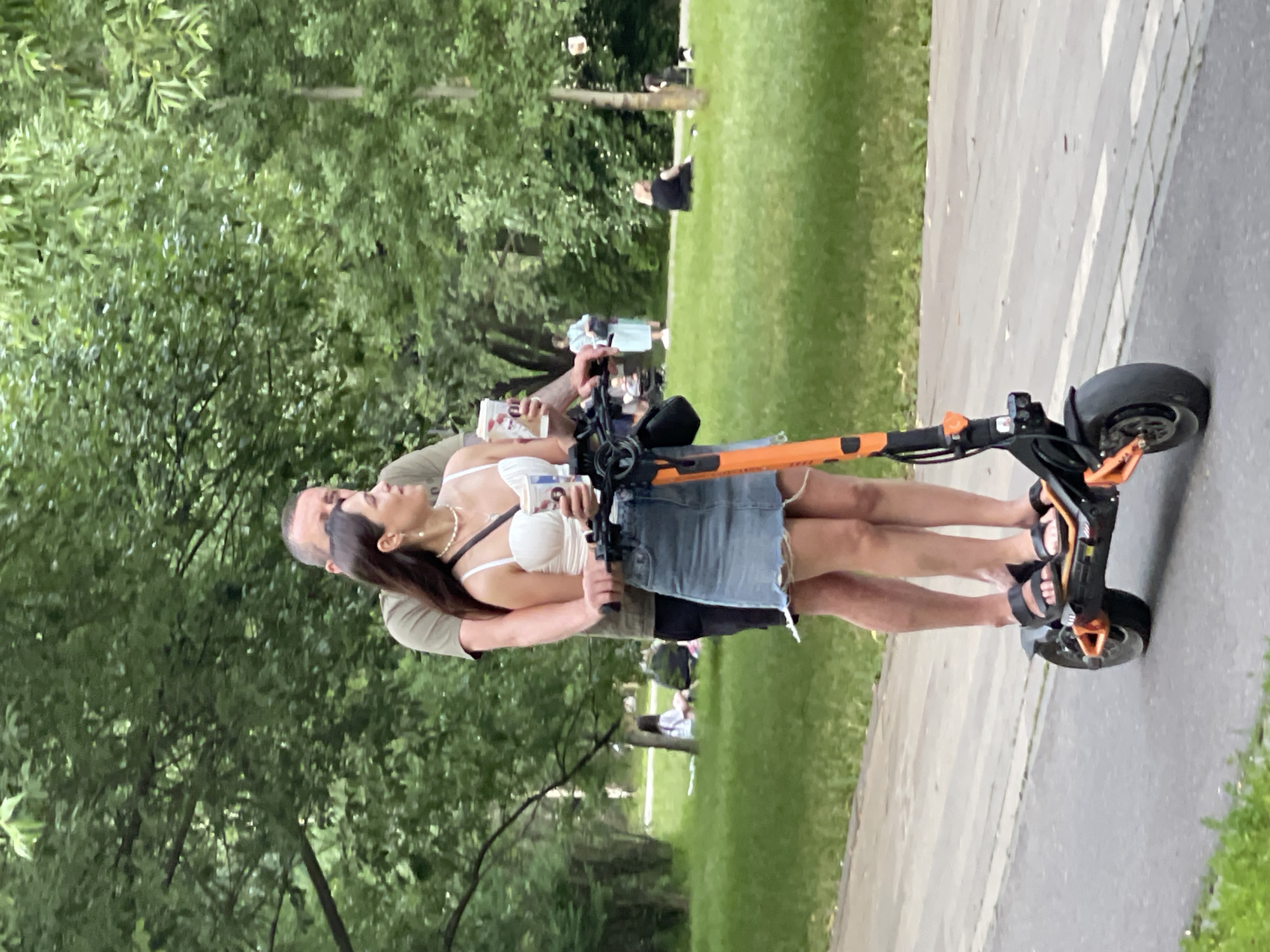 The image shows a man and a woman riding an orange electric scooter side-by-side on a paved path in a park. The man is standing behind the woman, with his arms around her. The woman is wearing a white top and a denim skirt, and the man is wearing a green t-shirt. Both are holding cups. In the background, there are trees, grass, and other people in the park. The context suggests this scene is likely in Gorky Park in Moscow, Russia, a popular urban green space known for its recreational activities.