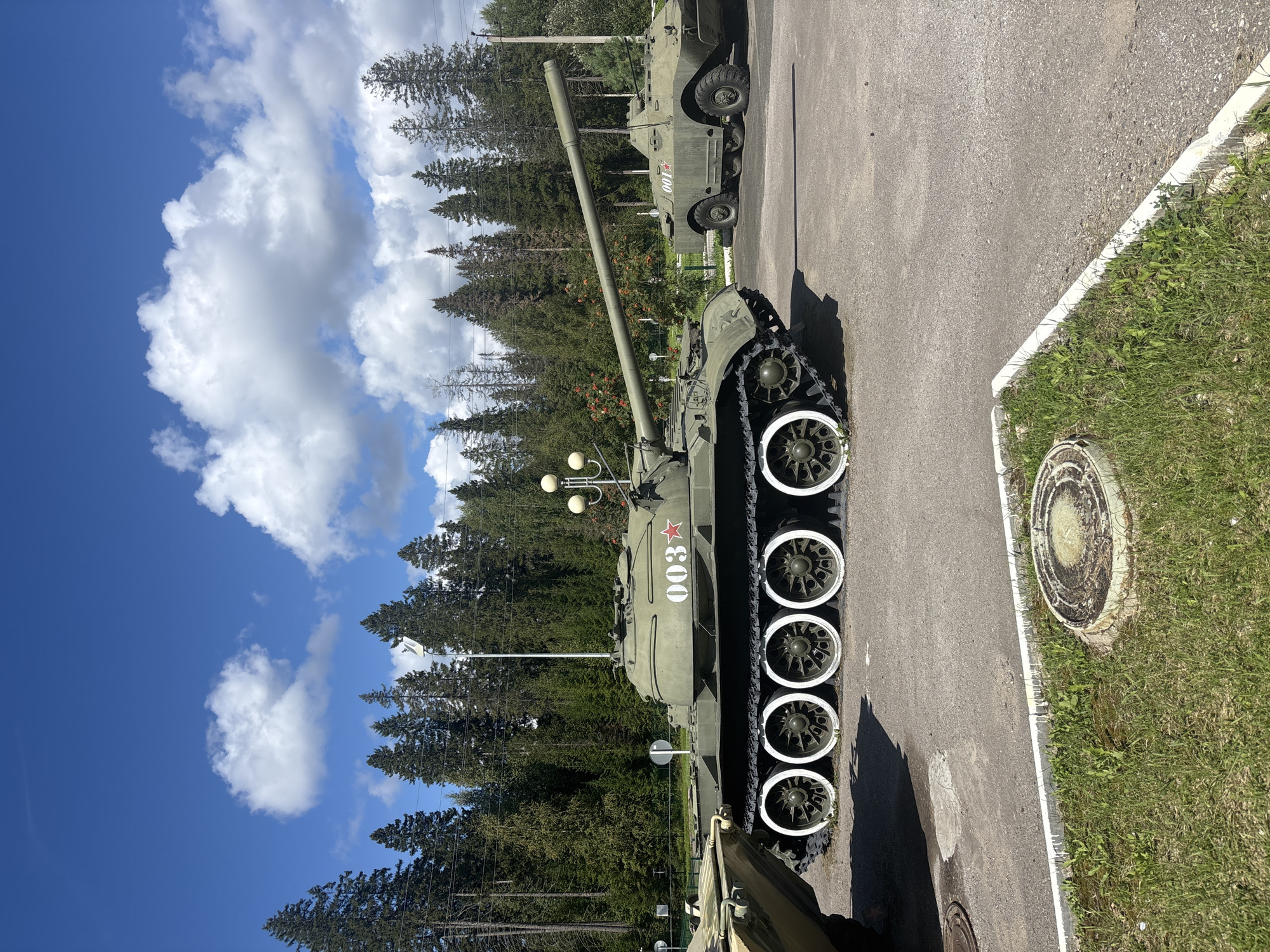 This image depicts an outdoor scene featuring military vehicles on a paved surface, with a dense forest and a bright blue sky with white clouds in the background. In the foreground, a green tank with the number "003" and a red star painted on its side is prominently displayed, featuring a long cannon and multiple white-rimmed road wheels. To the right of the tank, a smaller green armored vehicle with the number "001" and a red star is visible. The environment appears to be a museum or memorial site, possibly located in or near the city of Gamy, Russia, given the context provided. The lighting suggests a sunny day. There are no people visible in the image.