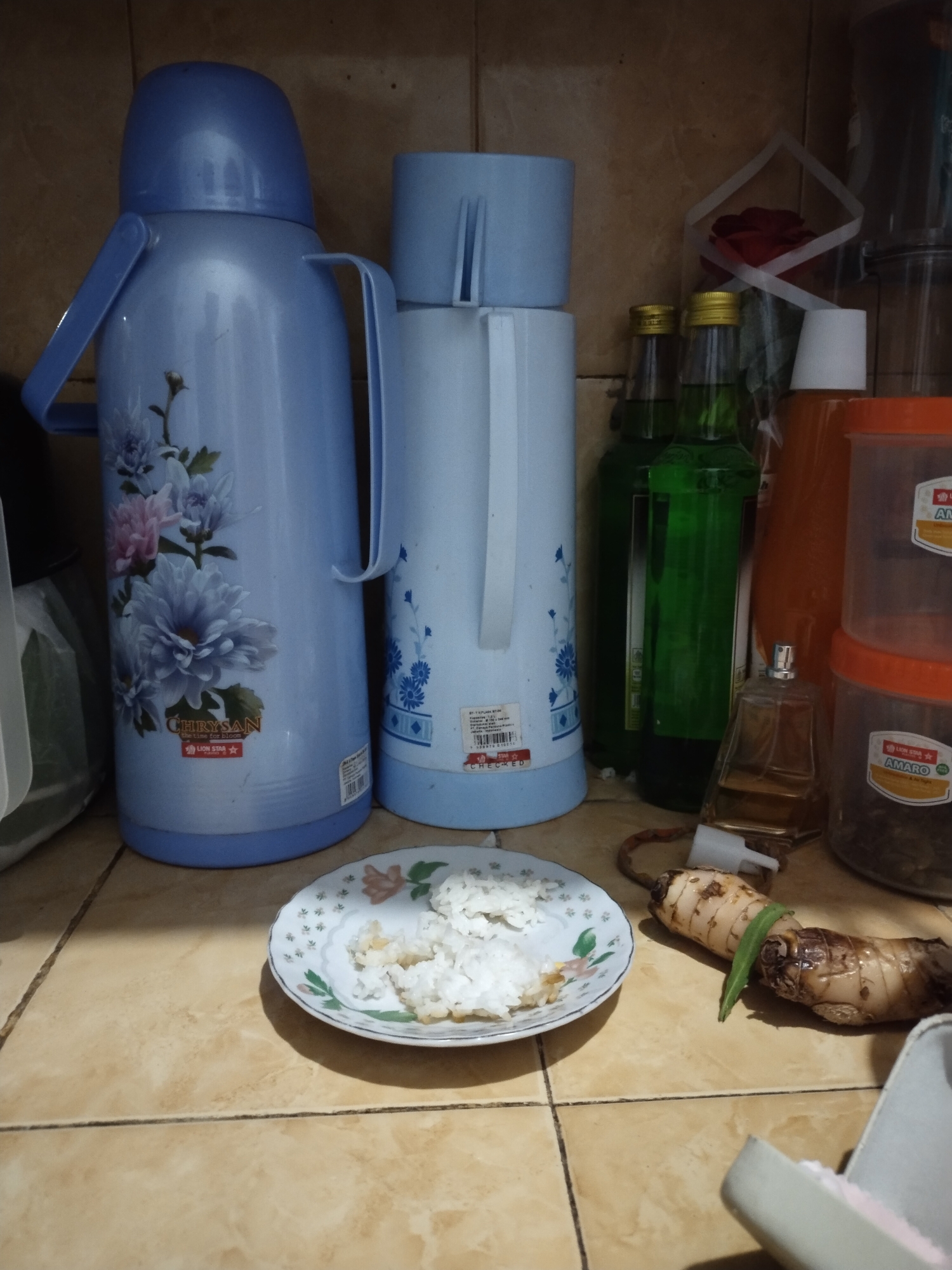 The image depicts a shelf in a kitchen or storage area, likely in Dermasandi, Indonesia. The shelf is made of tiled material, with brown and beige tiles.

Prominently displayed are two large light blue thermos flasks, one on the left and another slightly smaller one to its right. The larger flask on the left features a floral design of white and pink chrysanthemums. Both flasks have blue lids and integrated handles.

In front of the thermoses, a small white plate holds a portion of white rice, with some scattered pieces of food. To the right of the plate, two pieces of galangal, a spice with a rough, brown skin and a single green stem or leaf attached to one piece, lie on the tiled surface.

Behind the thermoses, several items are visible. Two green glass bottles with gold caps stand side-by-side. Further to the right, a clear glass perfume bottle with a golden liquid sits. Stacked transparent plastic containers with orange lids are also present on the right side of the shelf. One of these containers appears to hold dark-colored contents, possibly spices or dried goods. A white cone-shaped object, potentially a lid or part of a dispenser, is also visible. A hint of a red rose in a small bouquet can be seen in the background, behind the green bottles, partially obscured by a white paper or ribbon. The overall impression is one of a domestic setting with food-related items and containers.