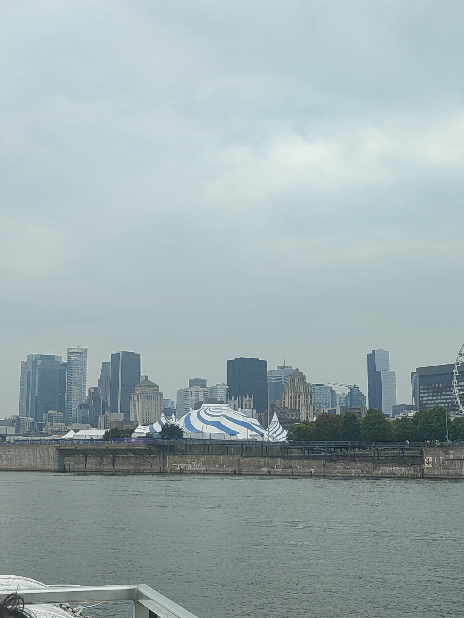 This media file captures a wide shot of the Montreal skyline on an overcast day. The scene is dominated by a dense collection of modern skyscrapers of varying heights and architectural styles, reflecting in the muted, grey sky. In the foreground, a body of water, likely the Saint Lawrence River, occupies the lower portion of the frame, with the edge of a boat visible in the bottom left corner.
Along the waterfront, a large, striped tent-like structure with blue and white patterns is prominent, suggesting an event or attraction. Behind it, a stone embankment with a dark metal railing forms a barrier between the water and the cityscape. The cityscape itself features a mix of glass-fronted towers and older, more ornate buildings. A Ferris wheel can be seen on the far right, another clue to a recreational area.
The weather appears to be cloudy, creating a diffused light that casts no harsh shadows and lends a slightly melancholic or subdued atmosphere to the image. The time of day is ambiguous due to the overcast conditions, but it seems to be daytime. The overall impression is of a bustling urban environment viewed from a distance, with a hint of leisure activity suggested by the tent and Ferris wheel. No discernible text is visible in the image.