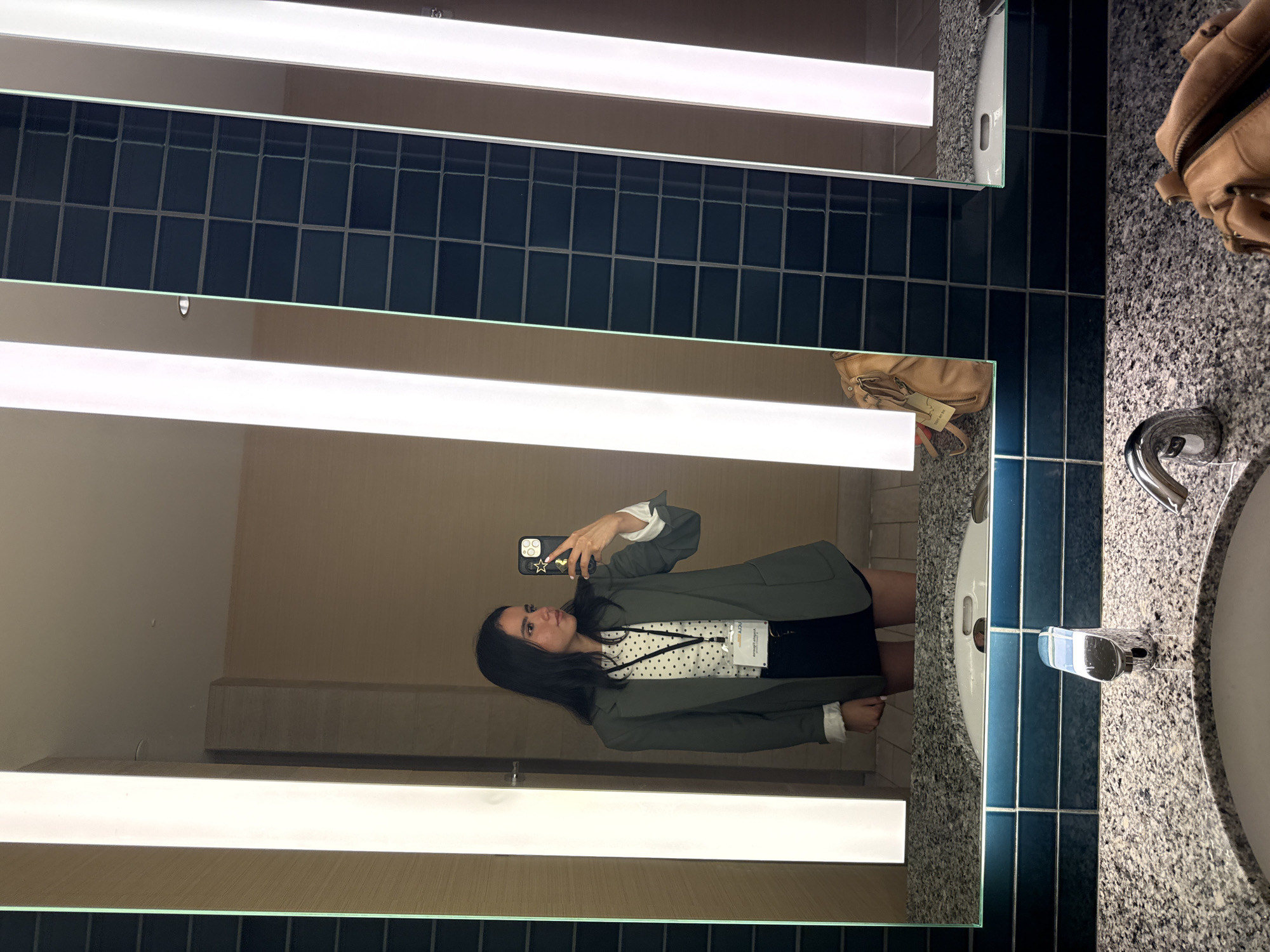 A young woman with dark hair is standing in a well-lit bathroom, taking a mirror selfie with her phone. She is wearing a green blazer over a white polka dot top and a black skirt, with a white lanyard around her neck. To her right, a tan leather handbag rests on the counter. The bathroom features a granite countertop with a modern faucet, and the wall behind the sink is tiled with dark blue squares. The overall environment appears to be a hotel bathroom. The scene is captured with the phone's front-facing camera.
