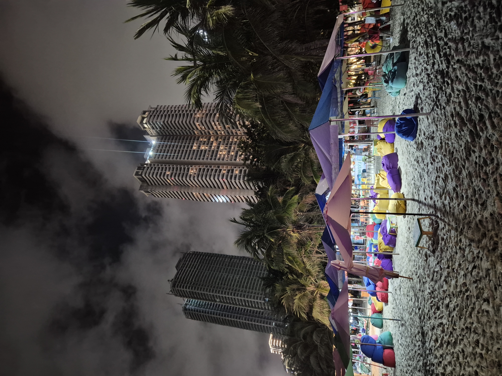 The image depicts a beach scene at night in Da Nang, Vietnam, with several tall, modern skyscrapers in the background. The beach is furnished with numerous colorful beanbag chairs arranged under a series of beach umbrellas, suggesting a recreational or dining area. Palm trees are visible between the umbrellas and the buildings, adding to the tropical ambiance. The sky is dark and cloudy, with some lights from the buildings illuminating the scene. The sand appears textured and slightly wet, typical of a beach at night. There are no people clearly visible in the foreground, though there may be figures in the distance under the umbrellas. The overall impression is a lively, illuminated beachfront area set against a backdrop of urban development.