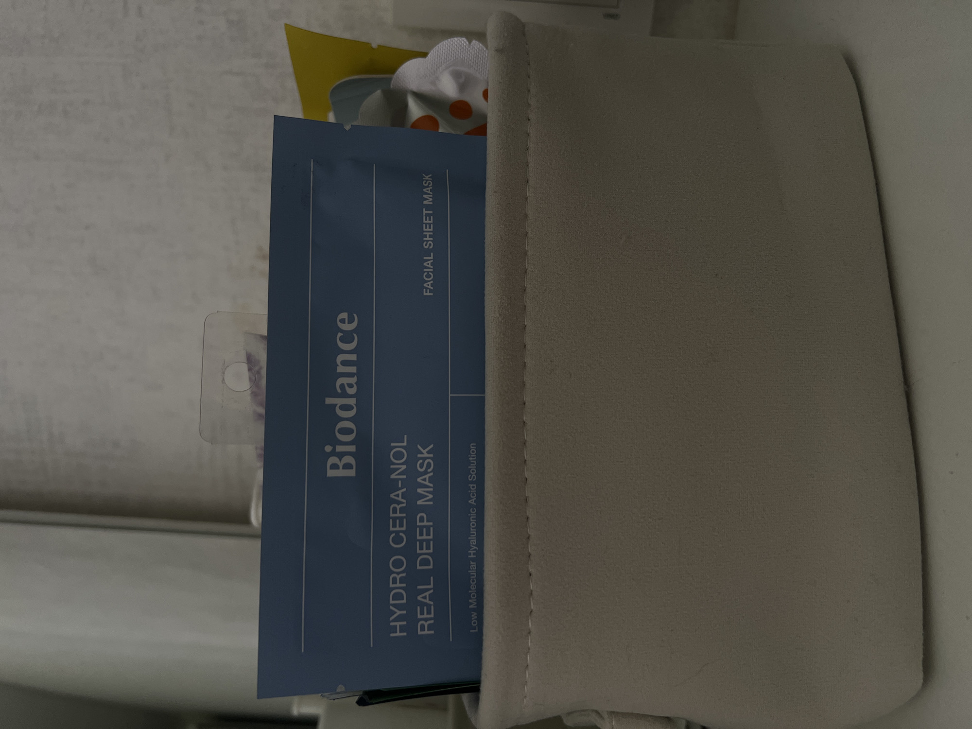 In an indoor, top-down shot, a light beige fabric basket contains a blue packet of Biodance Hydro Cera-Nol Real Deep Mask. The mask packet is angled and partially visible, with the brand name and product details clearly legible. The basket also holds other items, including a yellow object and a white object with orange dots. The background consists of a light-colored, textured surface and a portion of a white vertical structure. The lighting is dim. The location is Unknown, Unknown.