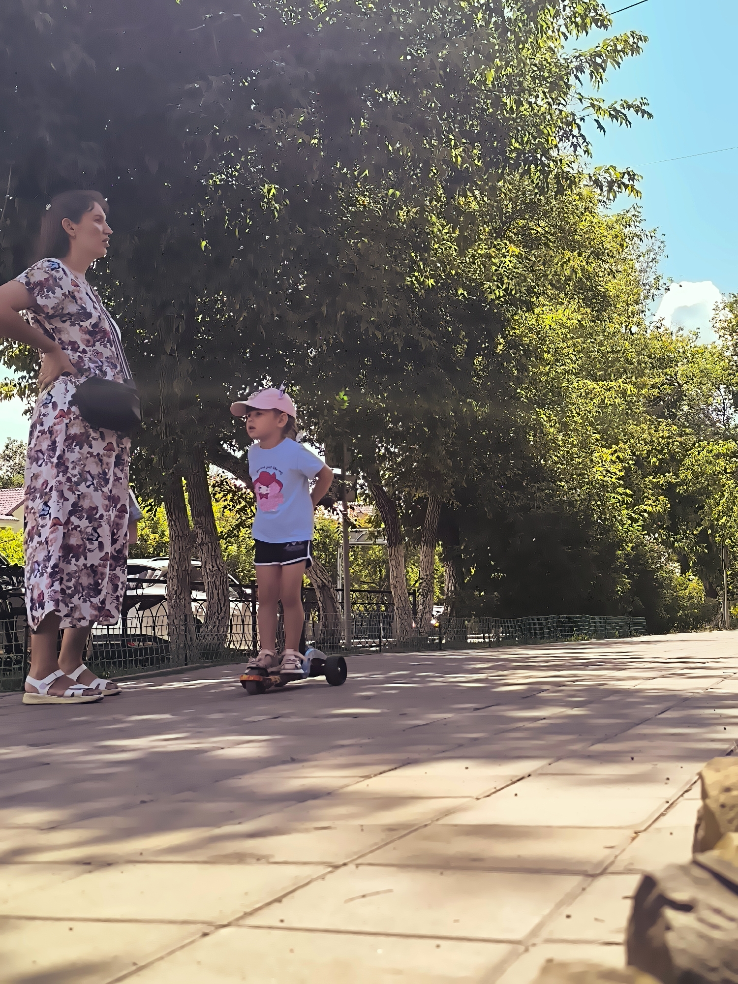 A woman stands with her hands on her hips, facing a young girl who is on a scooter. The woman wears a floral dress and sandals. The girl wears a pink cap, a blue t-shirt, and shorts. They are on a paved walkway in an outdoor setting with trees and a blue sky visible. The scene appears to be in a park or recreational area, possibly within the city of Karaganda, Kazakhstan, given the context provided.
