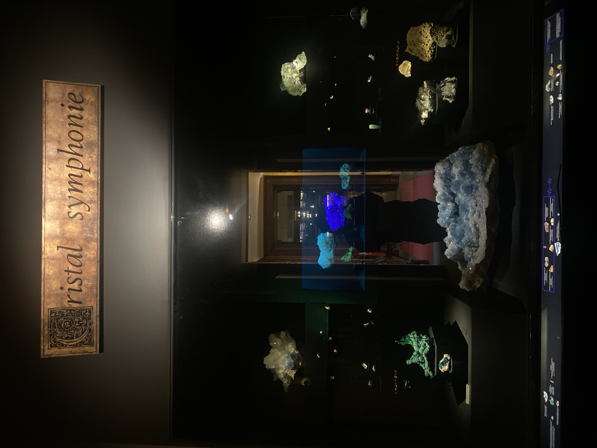 The image shows an exhibit titled "Cristal Symphonie" in a museum, most likely the Grenoble Natural History Museum, in Grenoble, France. The exhibit features a display case filled with various mineral specimens, some of which are illuminated with colored lights (blue and green). The display case has an interactive information panel on the right side. Through the back of the display, one can glimpse into another room. The foreground shows the wall and the exhibit sign.
