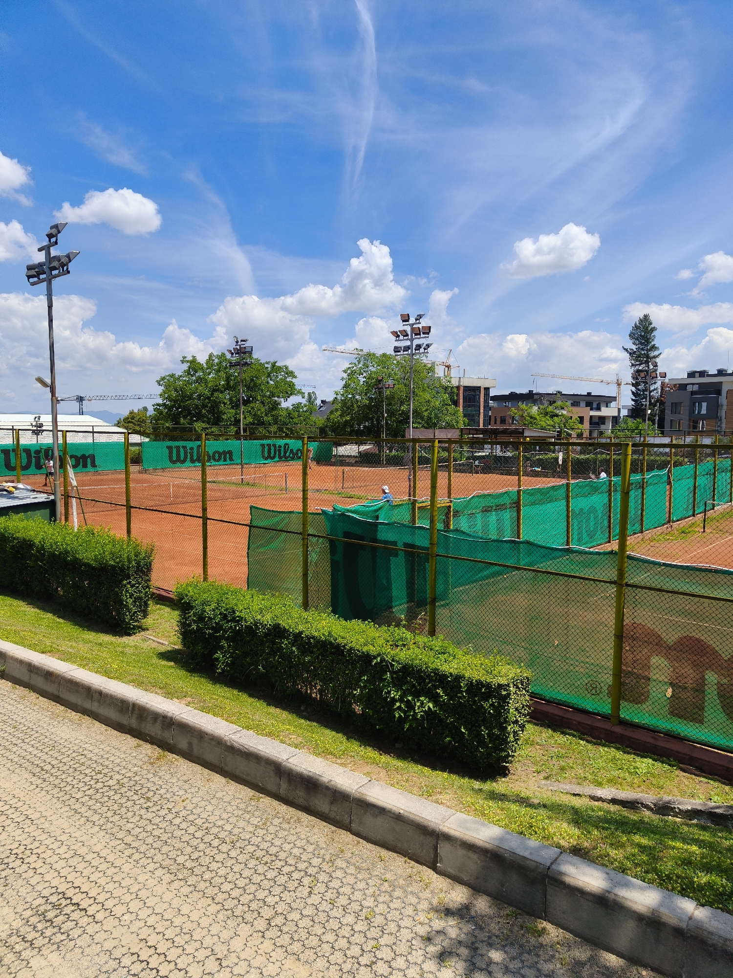 This image depicts an outdoor clay tennis court with a perimeter fence and green netting on the exterior. The court is equipped with floodlights and surrounded by manicured hedges and grass. In the background, there are modern buildings and construction cranes, suggesting an urban setting. Two individuals are visible on the court, seemingly engaged in playing tennis. The sky is blue with scattered white clouds, indicating fair weather. The presence of "Wilson" branding on the court's netting suggests it is a facility associated with the tennis brand. Considering the context of the surrounding environment and architectural style, this location could potentially be within the city of Sofia, Bulgaria, a region experiencing significant urban development.