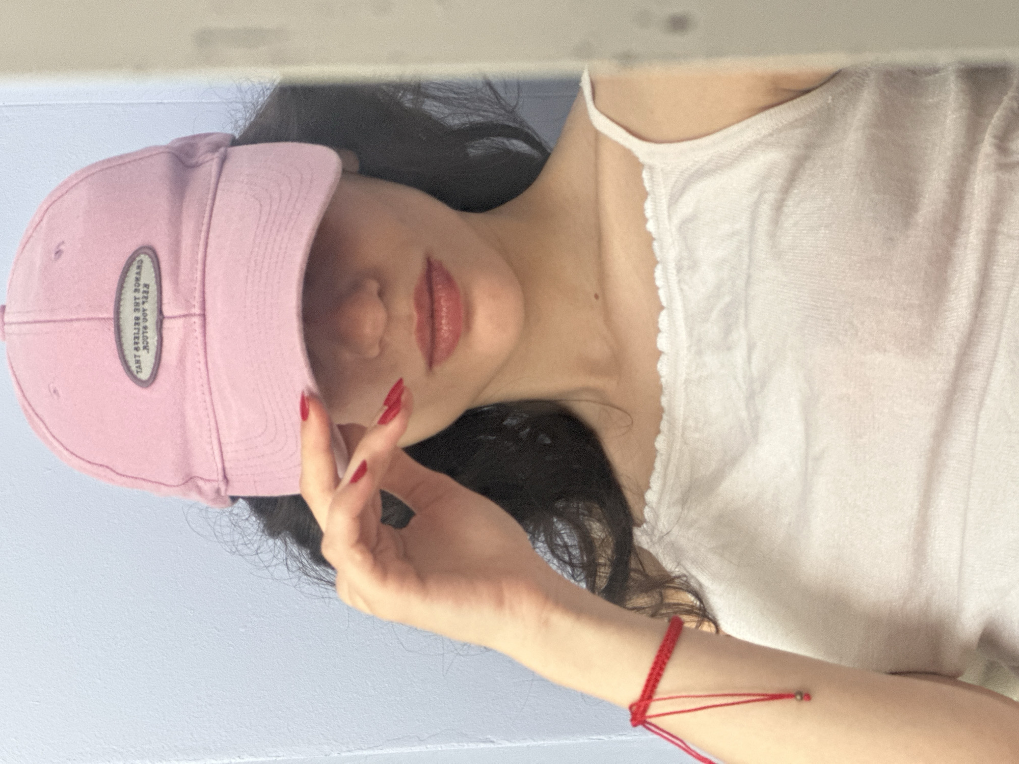 The image shows a young woman wearing a pink baseball cap and a white tank top. She is holding her hand up to her face, partially obscuring it. A red string bracelet is visible on her left wrist. The background appears to be a light blue wall. The woman is looking into a mirror, as a reflection of her right side is visible. The location is not discernible from the image.