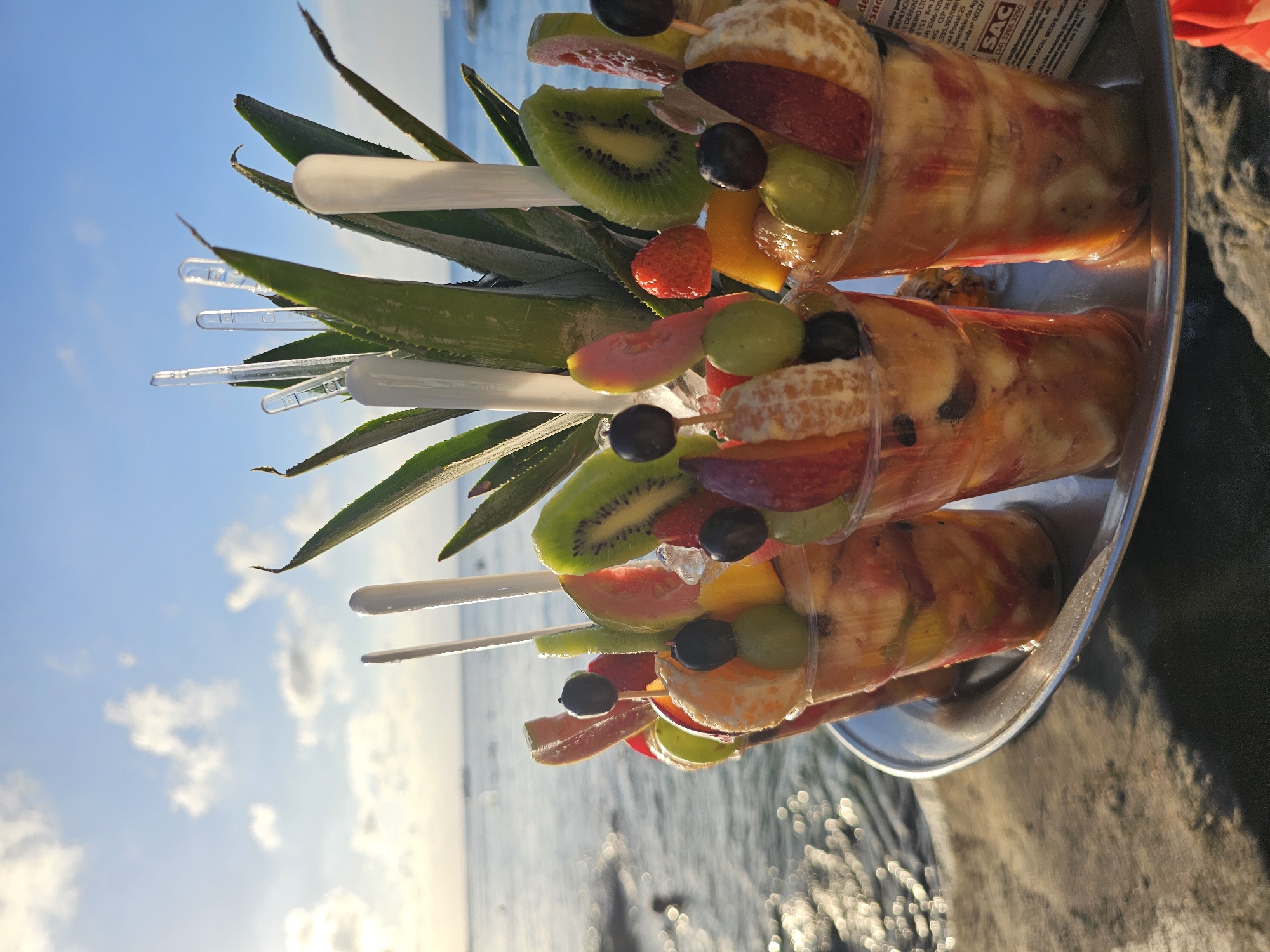 The image displays three plastic cups filled with fruit salad, arranged on a silver tray with pineapple leaves and plastic skewers sticking out. The fruit salad includes kiwi, strawberries, grapes, oranges, guava, and other unidentified fruits. The scene is set against a backdrop of the ocean and a partly cloudy sky. The cups appear to be sitting near the beach. Some packaging is visible. Based on these clues, the location is Armação dos Búzios, Brazil.