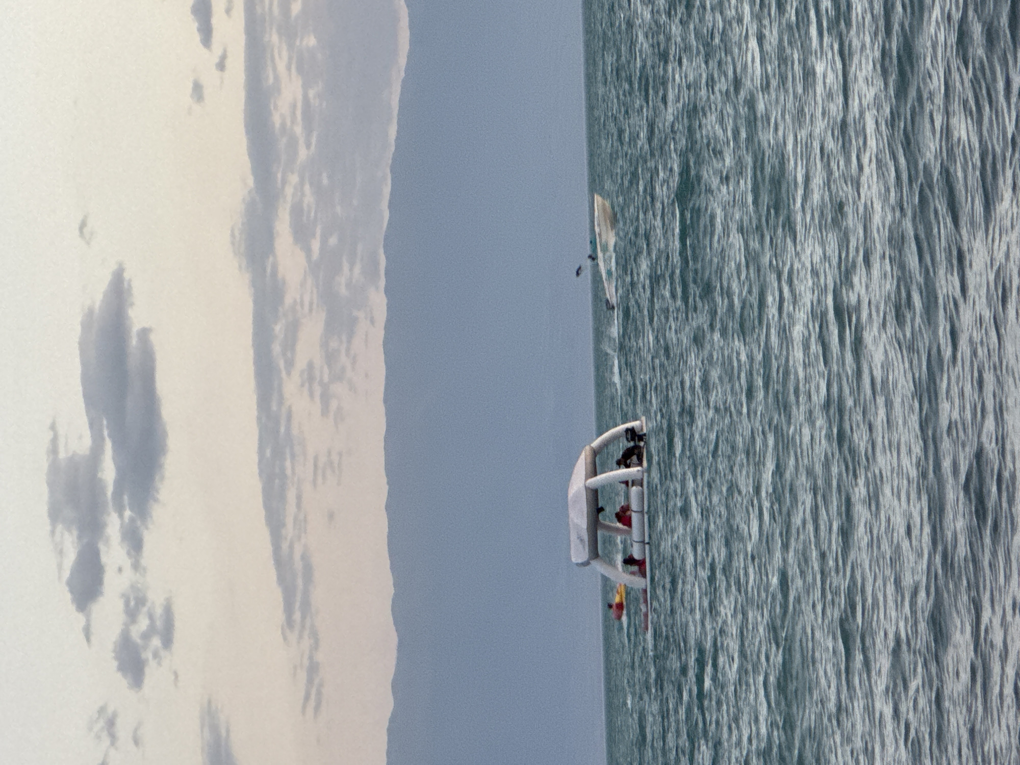In the city of Zharbulak, Kazakhstan, two boats are visible on the water. The first boat, a white inflatable raft with a canopy and several people aboard, is positioned in the foreground. To the left of this raft, a yellow inflatable tube is also on the water with a person on it. Further in the distance, a white speed boat is cutting through the water, creating a wake. The background features a mountain range under a cloudy sky, suggesting it is either early morning or late evening. The water appears choppy, indicating a breezy condition.