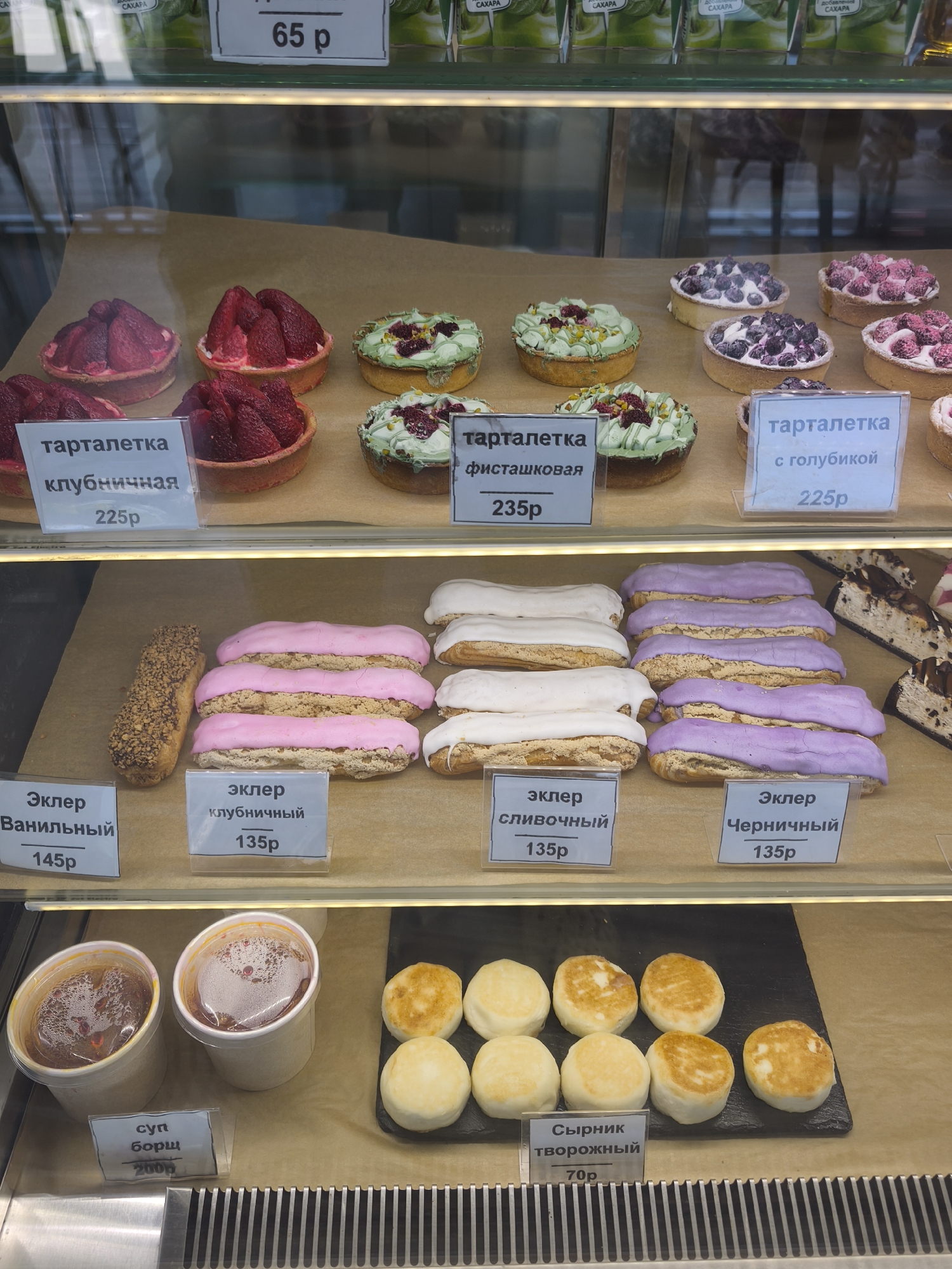 A display case in a food establishment shows a variety of desserts and some soup. On the top shelf, there are several tarts, including strawberry tarts, pistachio tarts with berries, and blueberry tarts. One sign indicates "65 p". On the middle shelf, there are eclairs of various flavors, including vanilla, strawberry, cream, and blueberry. On the bottom shelf, two containers of borscht soup are visible, with a sign indicating "суп борщ 200p". Next to the soup are several syrniki (cheese pancakes), with a sign indicating "сырник творожный 70p". The display case appears to be well-lit, and the food items are arranged neatly. There are no people visible in the image.