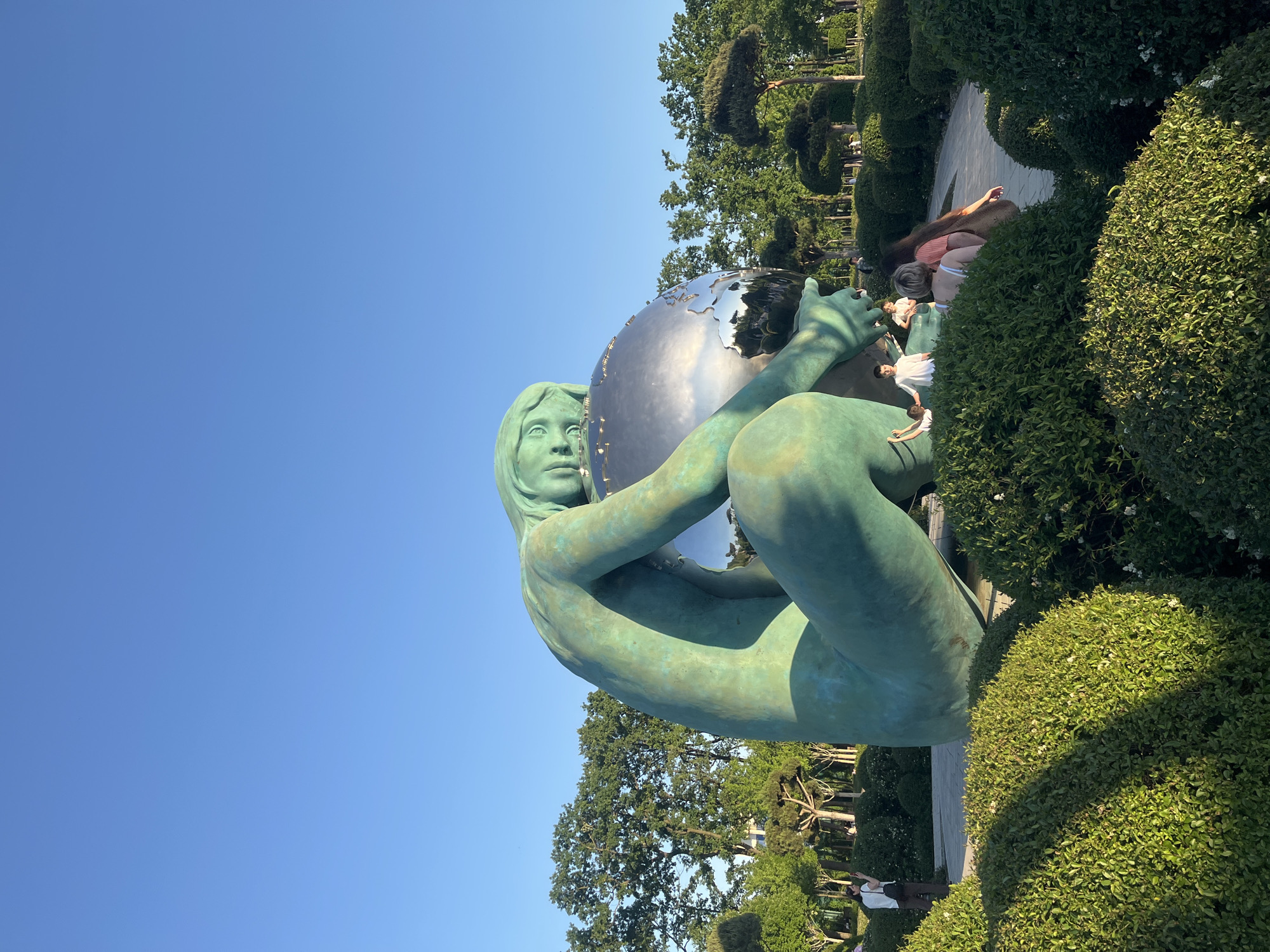 The image captures a monumental outdoor sculpture situated in a sun-drenched public park, identified as being in Krasnodar, Russia. The central subject is a large, teal-colored statue of a seated or crouching female figure. The sculpture possesses a smooth, verdigris-like patina, suggesting it is crafted from bronze or a similar material. The female figure is depicted cradling a large, highly reflective, metallic sphere, which appears to represent a globe or the Earth. Her face is serene, with her gaze directed slightly upwards and outwards, conveying a sense of contemplation or guardianship over the sphere.
The background features a clear, bright blue sky with no visible clouds, indicating excellent weather conditions. Surrounding the sculpture is a meticulously landscaped environment comprising lush, well-maintained green hedges and mature trees. Some trees in the upper right background display distinctive, almost topiary-like shapes. Several individuals are visible in the distance, casually walking or standing amongst the greenery, suggesting a peaceful day at the park. The strong sunlight and clear skies indicate it is daytime, likely late morning or early afternoon. No discernible text is visible within the frame. The overall scene portrays a harmonious blend of art, nature, and public leisure.