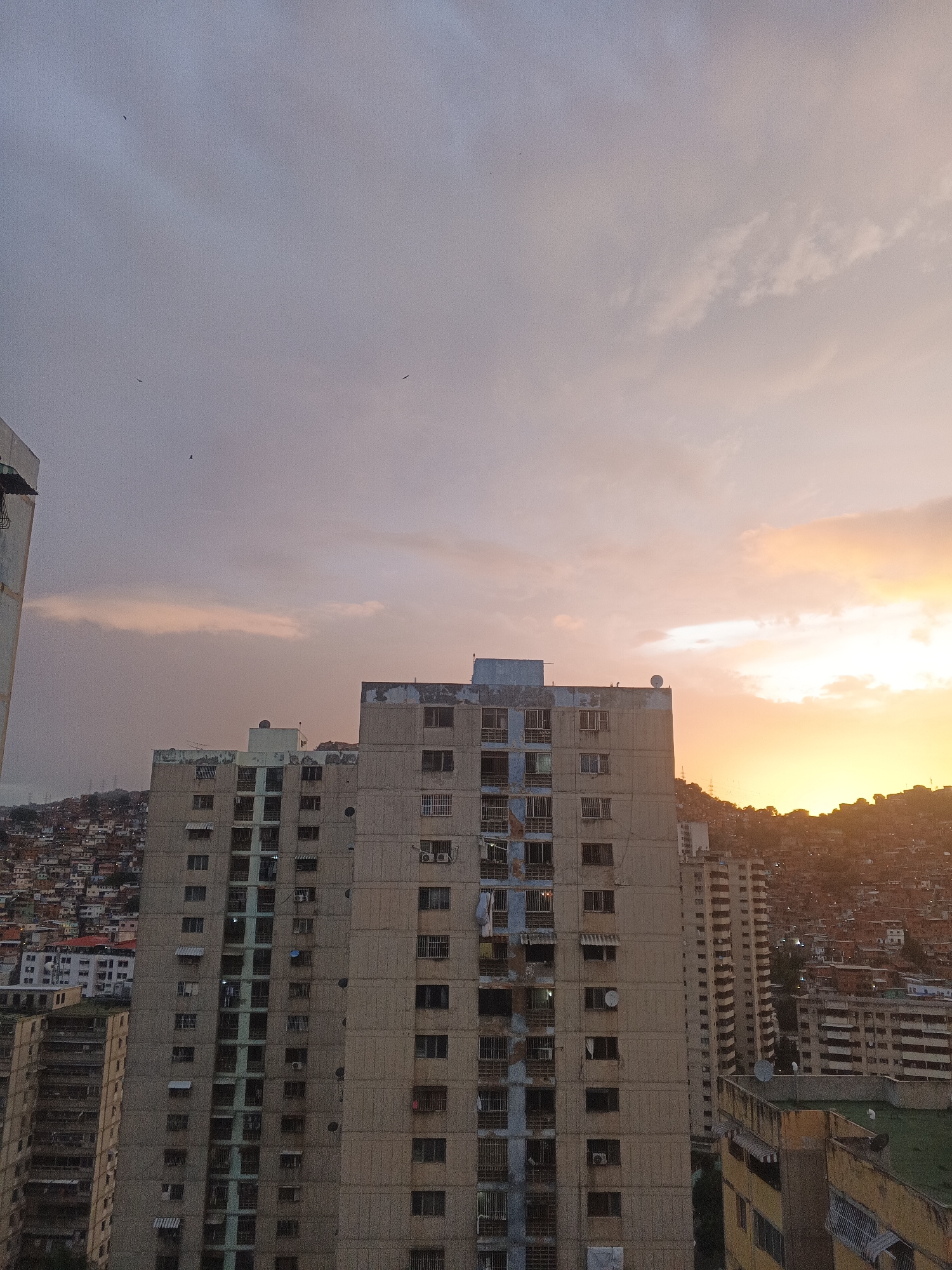 The image captures a view of the city of Caracas, Venezuela, at dusk. In the foreground, a tall, beige apartment building with multiple stories and numerous windows dominates the center of the frame. The building shows signs of wear and tear, with some windows boarded up or appearing faded. To the left of this building, another, lighter-colored building is partially visible. Behind the main apartment building, a dense hillside neighborhood with smaller, densely packed houses is visible, characteristic of Caracas's informal settlements, often referred to as "barrios."
The sky is filled with dramatic clouds, tinged with the warm hues of orange and yellow from the setting sun, creating a gradient effect towards the lighter, purplish-gray clouds on the left. Several birds are seen in silhouette against the sky. In the background, the silhouette of the surrounding hills can be seen, with more buildings scattered across their slopes. The overall scene suggests an urban environment with a strong contrast between the imposing apartment blocks and the sprawling informal housing on the hillsides, all set against a vibrant sunset.
