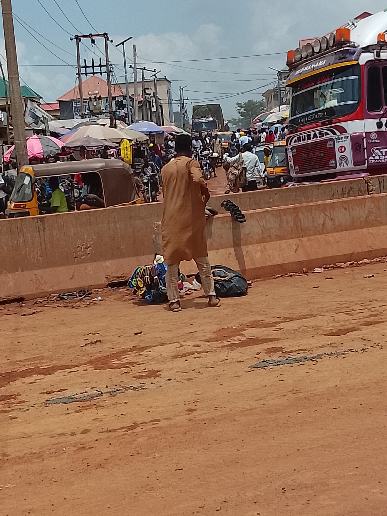 The image shows a street scene in Ado, Nigeria, characterized by a busy market area with a variety of transportation methods present. In the foreground, a man wearing a brown caftan and light-colored pants stands next to a low concrete barrier. He is surrounded by his belongings, including bags and what appears to be footwear. The ground is reddish-brown and uneven.
Behind the barrier, there is a congested market area with people, auto rickshaws (tuk-tuks), and motorcycles. Colorful umbrellas shade vendors and customers.
A large, decorated truck, with the words "ANUBAS," "MS-MAN-AMA," and "follow me transport" visible on its front, is positioned to the right of the market area. The truck's license plate reads "CH 17-232." Power lines and utility poles rise above the buildings in the background. In the distance, other vehicles and structures are visible.
