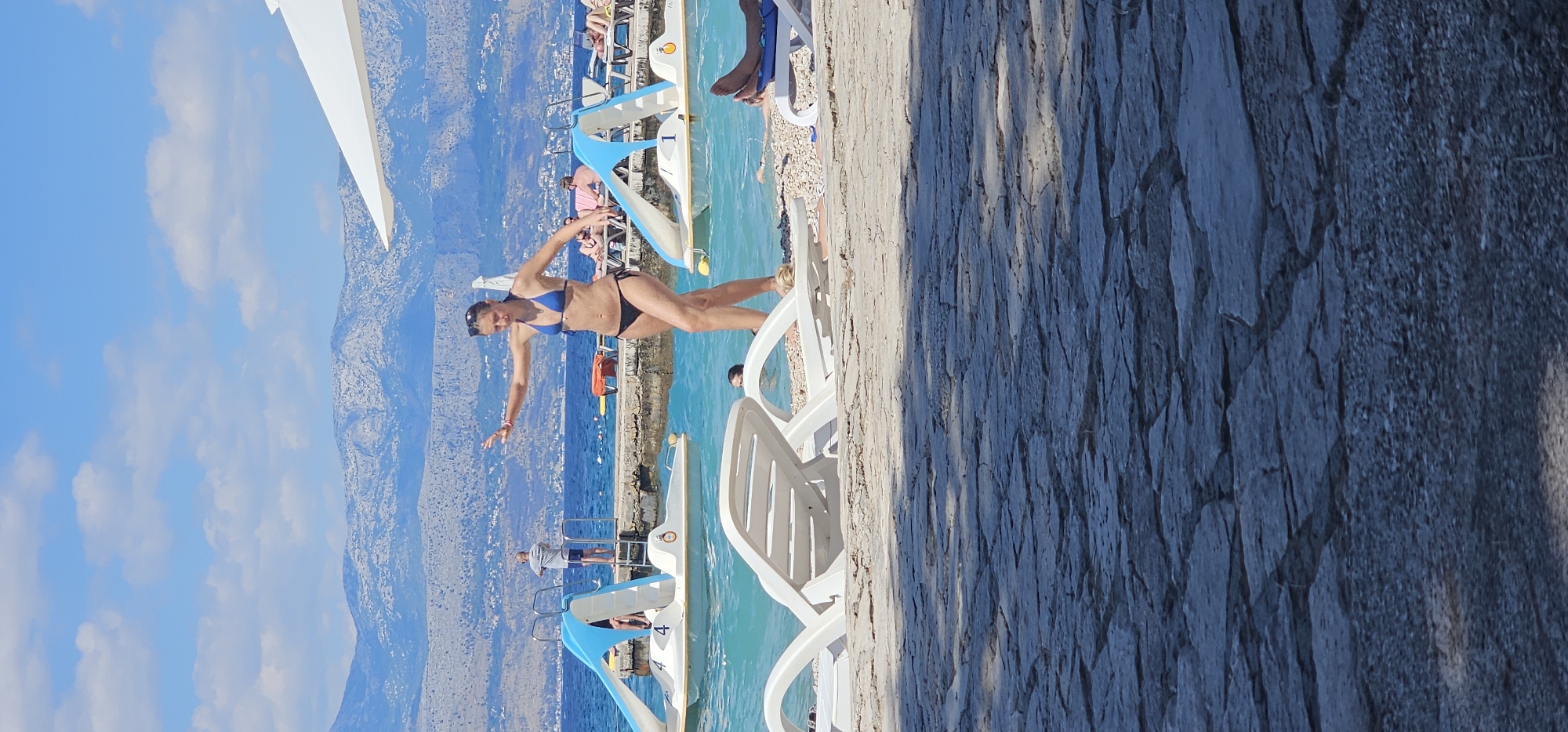 In the city of Supetar, Croatia, on a sunny day, a woman in a blue and black bikini stands on a pebble beach, her arms outstretched. The turquoise water laps at the shore, where several white lounge chairs are placed. In the background, a pier extends into the sea, with a man standing on it and two paddle boats moored nearby. The scene is framed by a hazy mountain range in the distance and a bright blue sky dotted with white clouds. The foreground is dominated by a textured stone pathway.