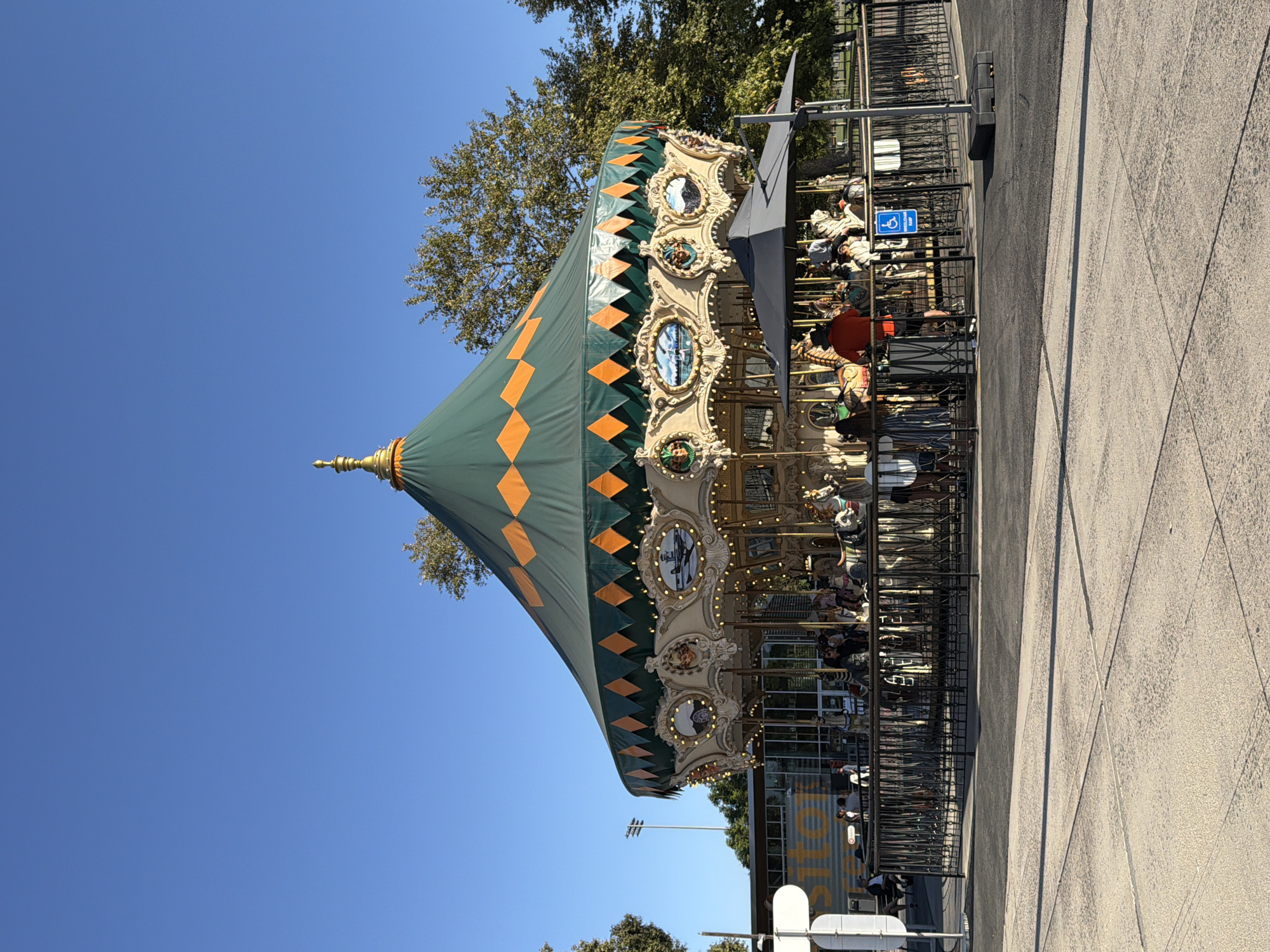 The image shows a carousel in a park on a clear, sunny day. The carousel is ornate, with a green and orange striped canopy and decorative elements around its perimeter. Several people are riding the carousel, and others are standing around the perimeter. A black metal fence surrounds the carousel, and there is a blue accessibility sign on the fence. In the background, there is a modern building with large windows, trees, and a clear blue sky. The ground in the foreground is paved with concrete. The scene is located in Irvine, California, United States of America.