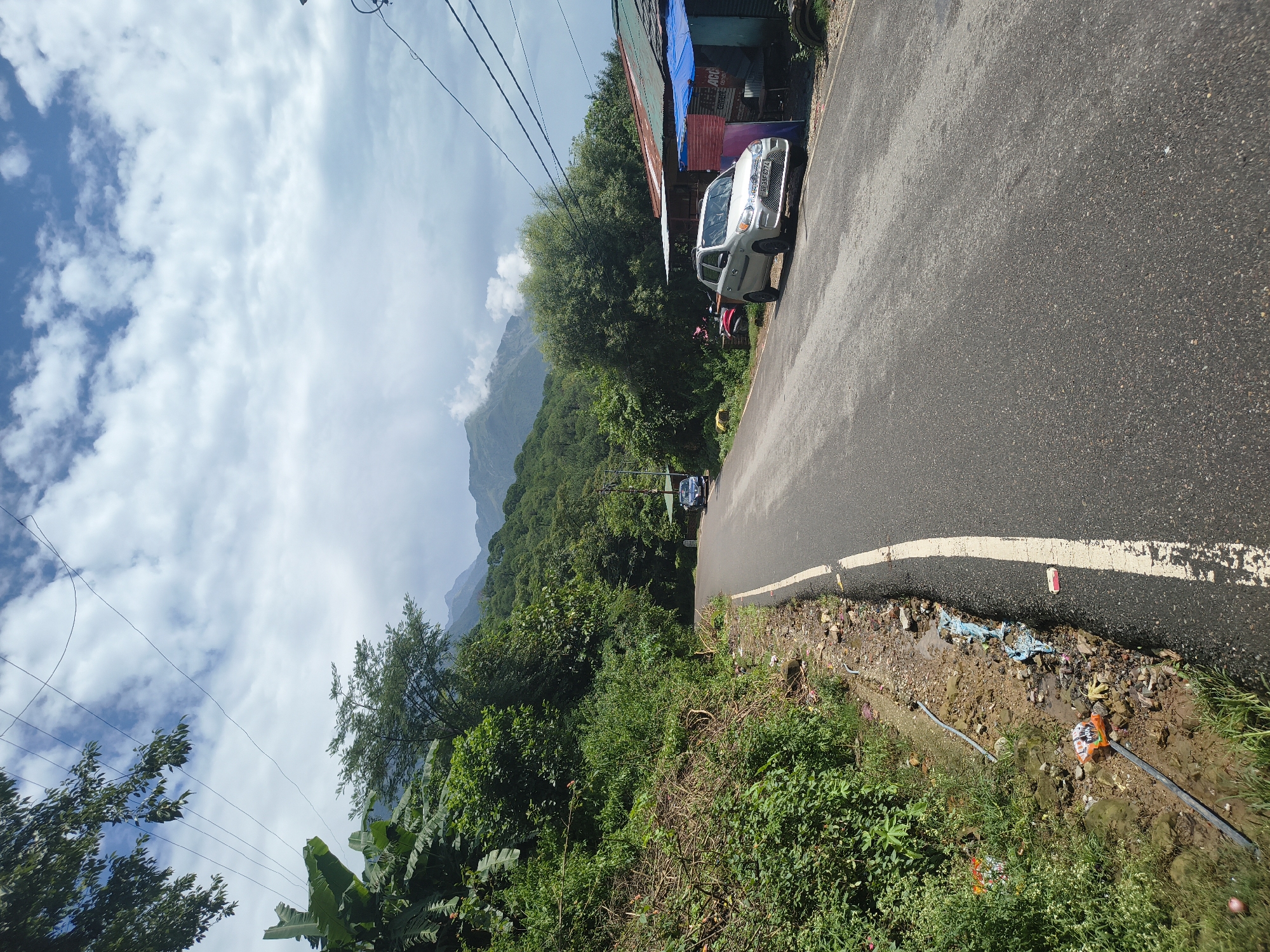 The image depicts a rural road curving through a lush green, mountainous landscape, likely in or near Kandi, India. The road is asphalt with a white line marking its edge. On the left, a steep embankment covered in dense vegetation, including banana plants, slopes upward. Debris, including plastic bags and wrappers, is visible along the roadside and in a small, muddy ditch.
On the right side of the road, a light-colored hatchback car is parked. Further down the road, a small roadside structure with a corrugated metal roof and blue tarpaulin is visible, with what appears to be a scooter parked beside it. In the background, rolling green hills and mountains rise under a partly cloudy sky. Power lines crisscross the upper portion of the image. The scene appears to be a snapshot of everyday life on a road in a scenic, underdeveloped area.
