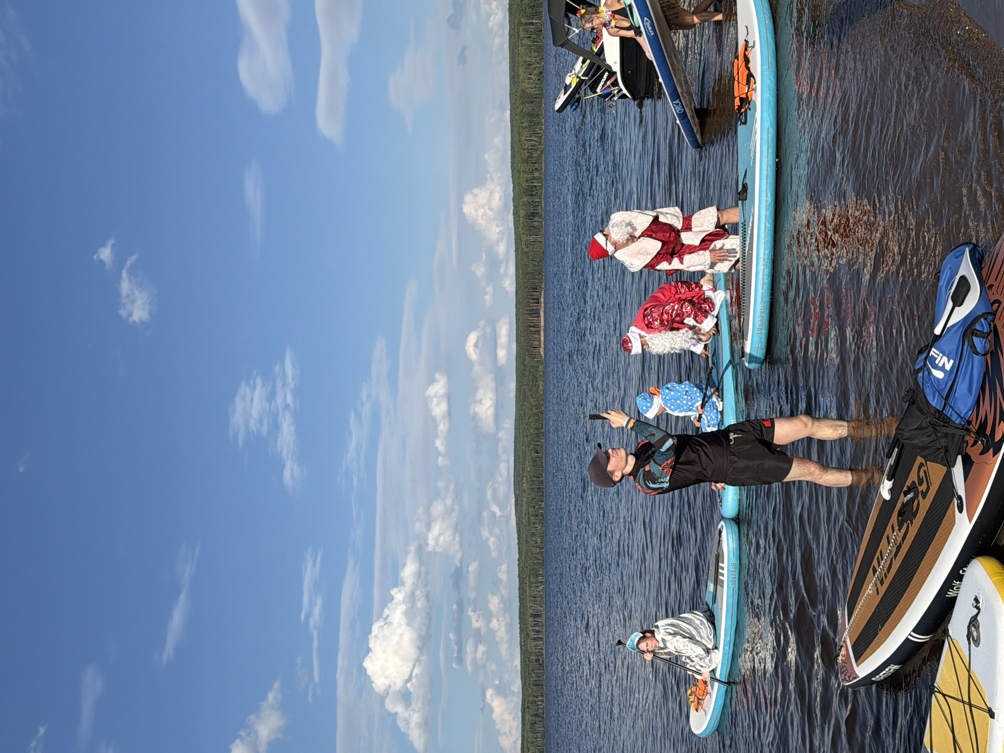 An outdoor, medium shot captures a group of people on paddleboards in a body of water, possibly a lake or bay, with a dense forest line visible in the background under a bright, cloudy sky. In the foreground, two paddleboards are partially submerged, one featuring a blue bag with "FIN" written on it. A person in a black long-sleeved shirt with a colorful, abstract pattern and black shorts stands in the water, holding a smartphone and looking at it. Next to them, a child wearing a blue snowsuit with a hood is seated on a paddleboard. Further back on blue paddleboards are three individuals dressed as Santa Claus, one in a red and white suit with a beard, another in a red suit with white trim and a beard, and a third Santa in a similar red and white outfit with a beard, kneeling. To the left, a person wearing a silver, hooded jacket sits on a paddleboard, holding a paddle. In the far right background, a portion of a boat with a black canopy is visible, with two people standing near it and on a paddleboard. The water is dark and rippled, reflecting the sky and surrounding environment. The scene appears to be set during daylight hours. There are no visible markers or landmarks to indicate the specific location in Novyi, Russia.