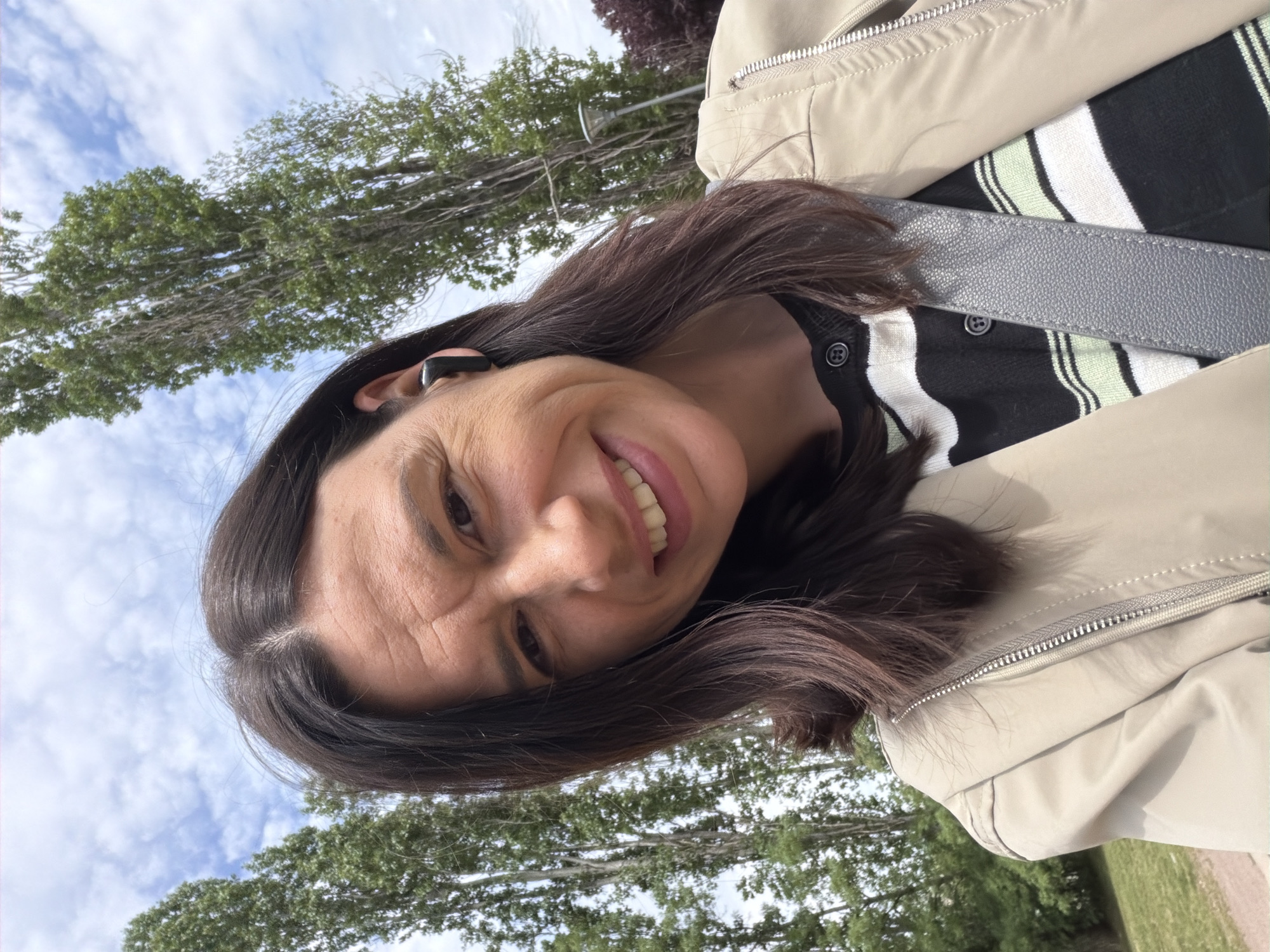 A woman with dark hair and a black earbud in her left ear is smiling and looking towards the camera. She is wearing a light beige jacket over a black and white striped cardigan. A gray shoulder strap is visible across her chest. The background shows trees and a cloudy blue sky, suggesting an outdoor setting. The photo was taken in Sofia, Bulgaria.