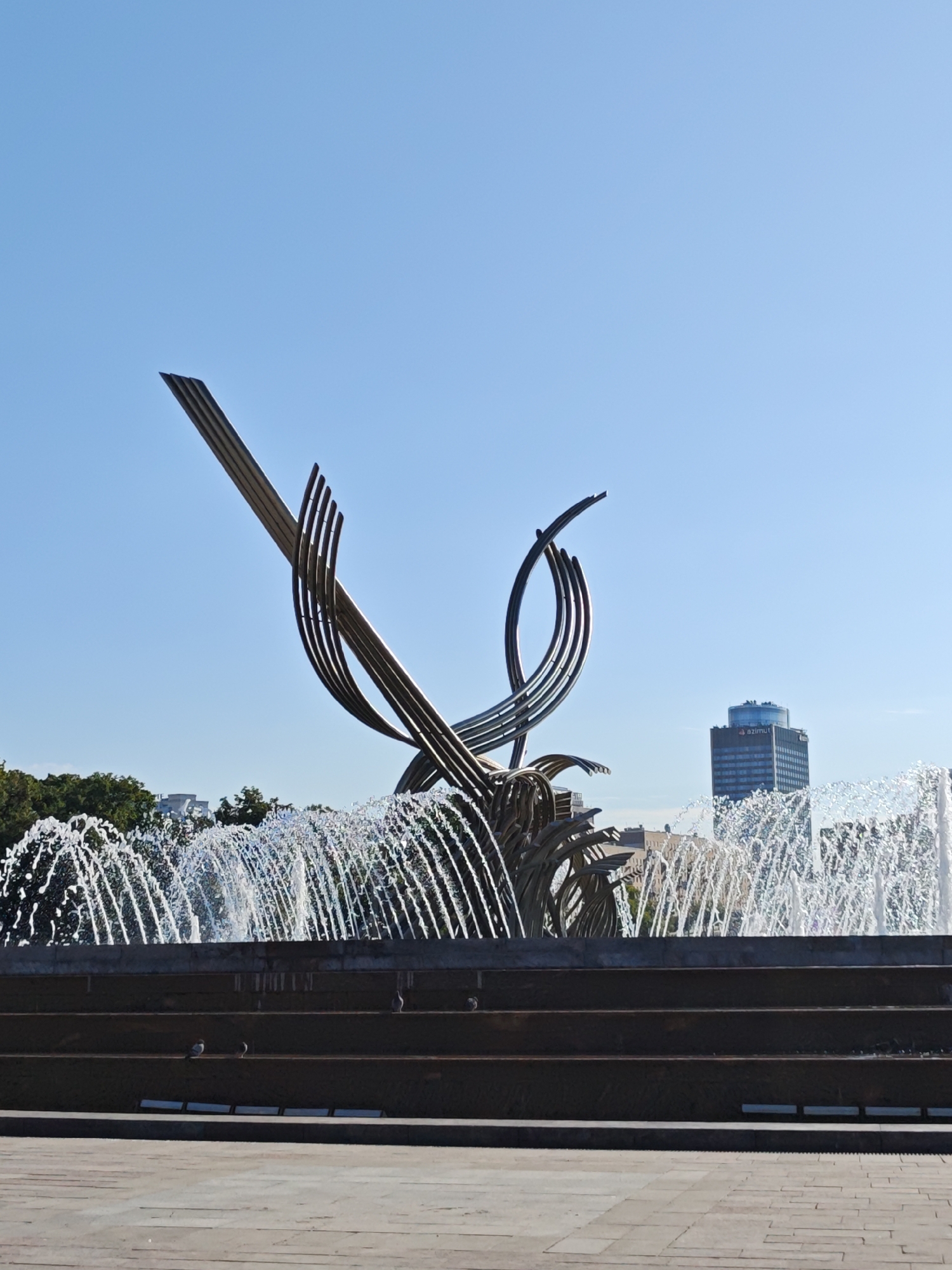 A large, abstract metal sculpture is the central focus of this image, set against a clear blue sky. The sculpture, made of curved and angled metallic elements, appears to be part of a fountain, as a continuous spray of water cascades in front of it. In the background, to the right of the sculpture, a modern high-rise building with a distinct rounded top and the word "azimut" visible on its facade stands out. Below the sculpture and fountain, tiered stone structures are visible, with several pigeons perched on the ledges. Trees line the horizon behind the fountain. The foreground consists of a paved area. The scene is likely located in a public space within the city of Moscow, Russia.