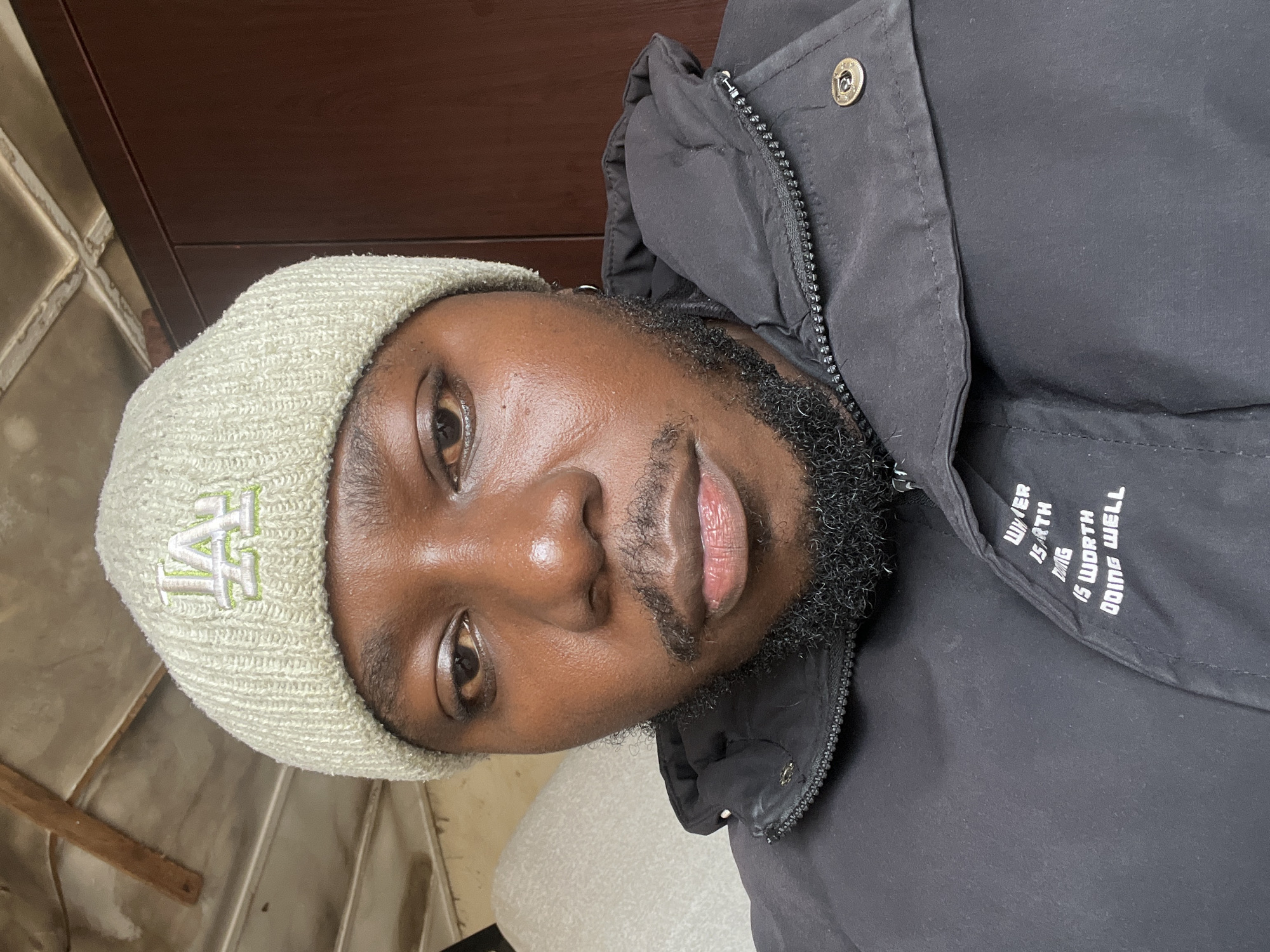 This is a medium close-up, selfie-style shot of a Black man in Jos, Nigeria. He is wearing a light green knitted beanie with a lime green "LA" logo embroidered on it, and a black puffer jacket with a zipper closure. His facial hair is short and dark. The background shows a wall and a piece of furniture. The lighting suggests an indoor setting, possibly during the day. The man's gaze is directed slightly downwards and to the left of the camera. The overall impression is casual and personal.