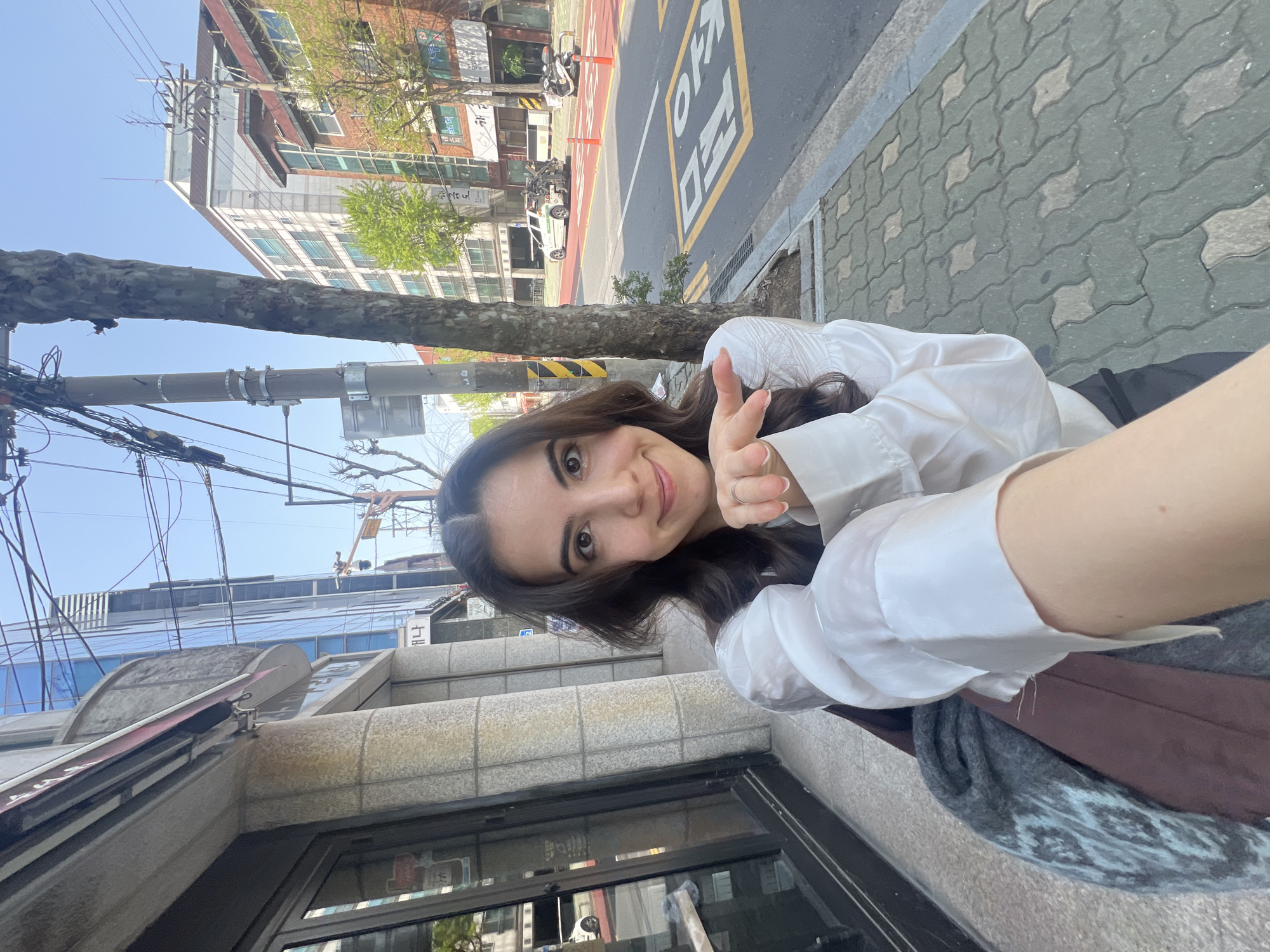 A young woman with long brown hair smiles and holds up two fingers in a peace sign while taking a selfie. She is wearing a white button-down shirt and appears to be seated, possibly on a stoop or step.
The setting is an urban environment, likely in Seoul, South Korea, as indicated by the Korean characters painted on the street pavement, "오지마". Behind her, there's a tiled sidewalk, a street with yellow painted markings, and buildings with Korean signage. Power lines crisscross the sky. The weather appears to be sunny and clear.