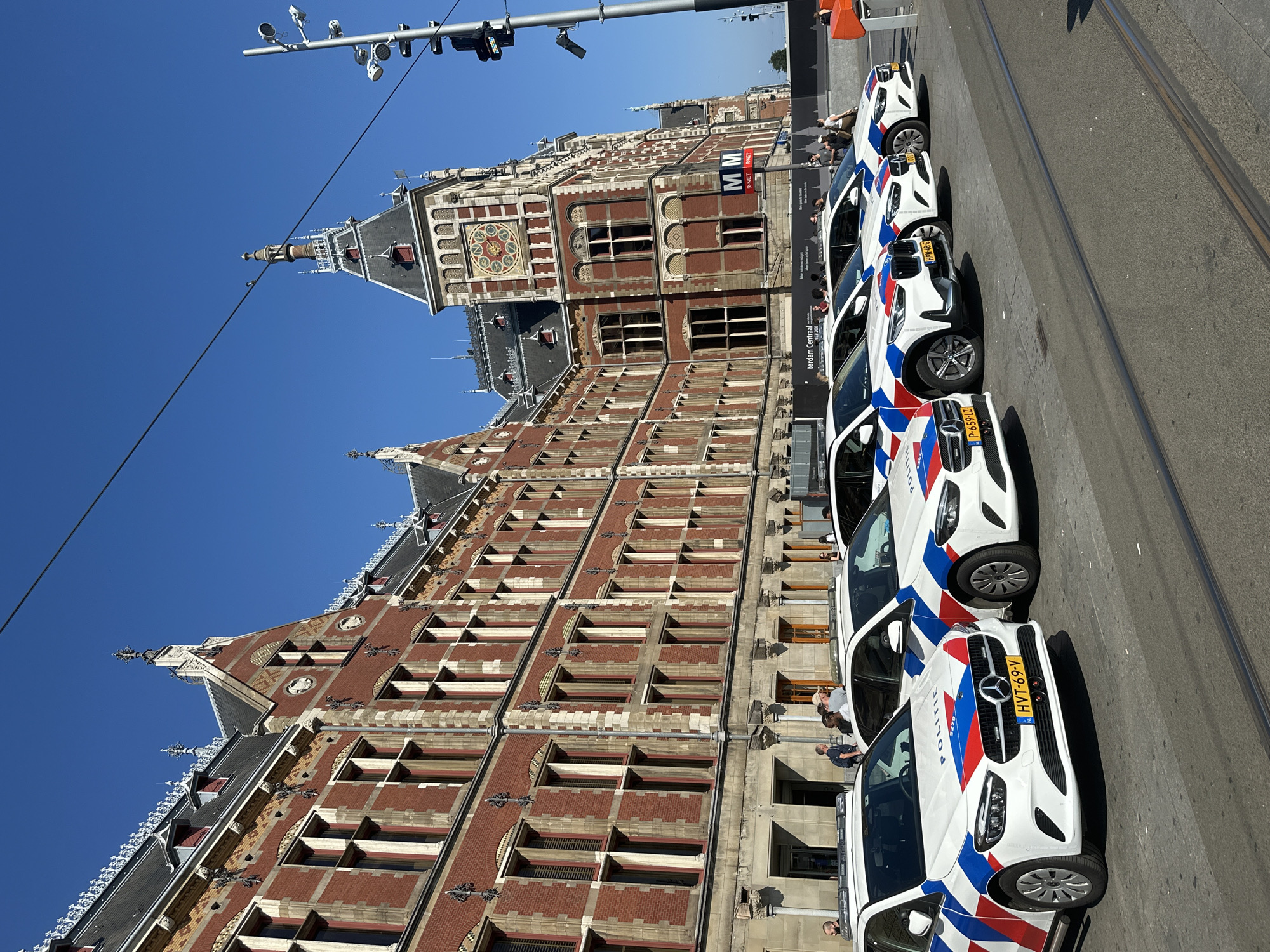 The image depicts a row of white police cars with Dutch "Politie" markings parked in front of a large, ornate red brick building. The building appears to be a historical landmark, possibly a train station, given the clock tower and grand architecture. The cars are modern Mercedes-Benz vehicles, equipped with light bars. Several people are visible in the background, some near the cars and others walking along the building. The scene is set on a sunny day, with a clear blue sky overhead. The presence of tram tracks in the foreground suggests the location is a public square or thoroughfare. Based on the architecture and the "Politie" markings, the location is identifiable as Amsterdam Centraal Station in Amsterdam, Netherlands.