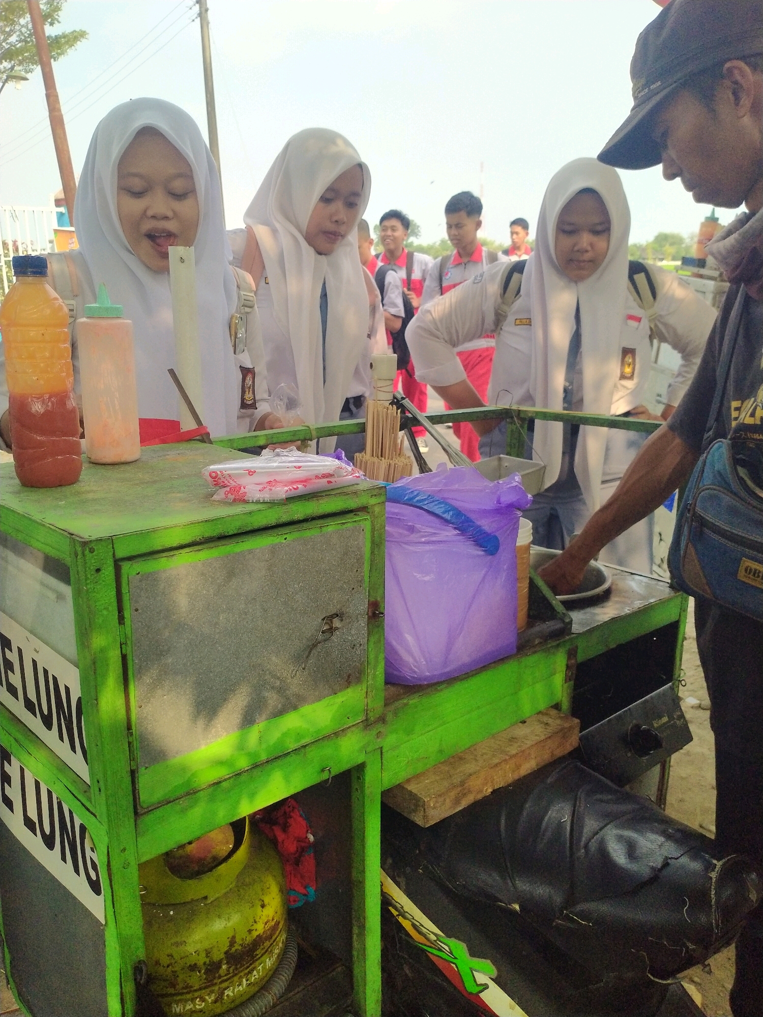 In Begajah, Indonesia, three female students wearing white hijabs and school uniforms are gathered around a green street food cart. One student on the left is leaning forward, her mouth open as if eating or speaking. Another student in the center is looking down, and the student on the right is interacting with a man wearing a dark baseball cap and a dark t-shirt. The man, who appears to be the vendor, is reaching towards the cart.
The street food cart is green and has a metal front panel. On top of the cart are two plastic bottles, one containing an orange liquid and the other a pinkish liquid, along with a stack of plastic-wrapped straws. A purple plastic bag and a small metal container are also visible on the cart. Behind the cart, other students in school uniforms can be seen in the background, along with trees and utility poles. A green LPG gas cylinder is visible on the lower shelf of the cart, partially covered by a red cloth. The ground appears to be dirt or gravel. The lighting suggests it is daytime, with a bright sky.