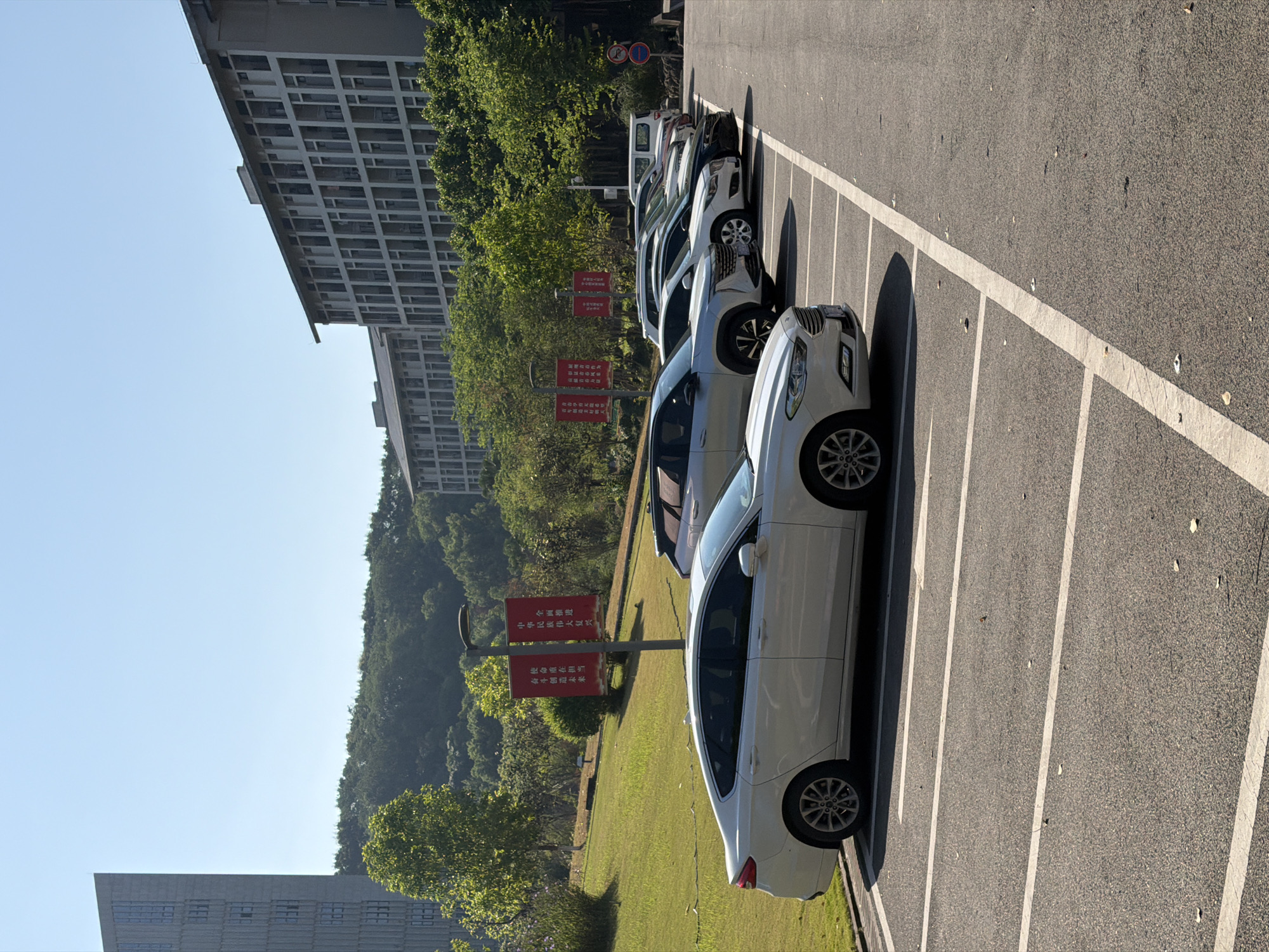 The image depicts a parking lot on a sunny day, likely in the city of Jiangjin, China. In the foreground, a white sedan is parked in a marked spot. Several other vehicles are parked in a row behind it, including SUVs and a white van. The parking lot is paved with asphalt and marked with white lines. In the background, there are several modern buildings with many windows, as well as lush green trees and rolling hills. Two red banners hang from poles in the middle ground, with Chinese characters visible on them. The sky is clear and blue. There are no people visible in the image.
