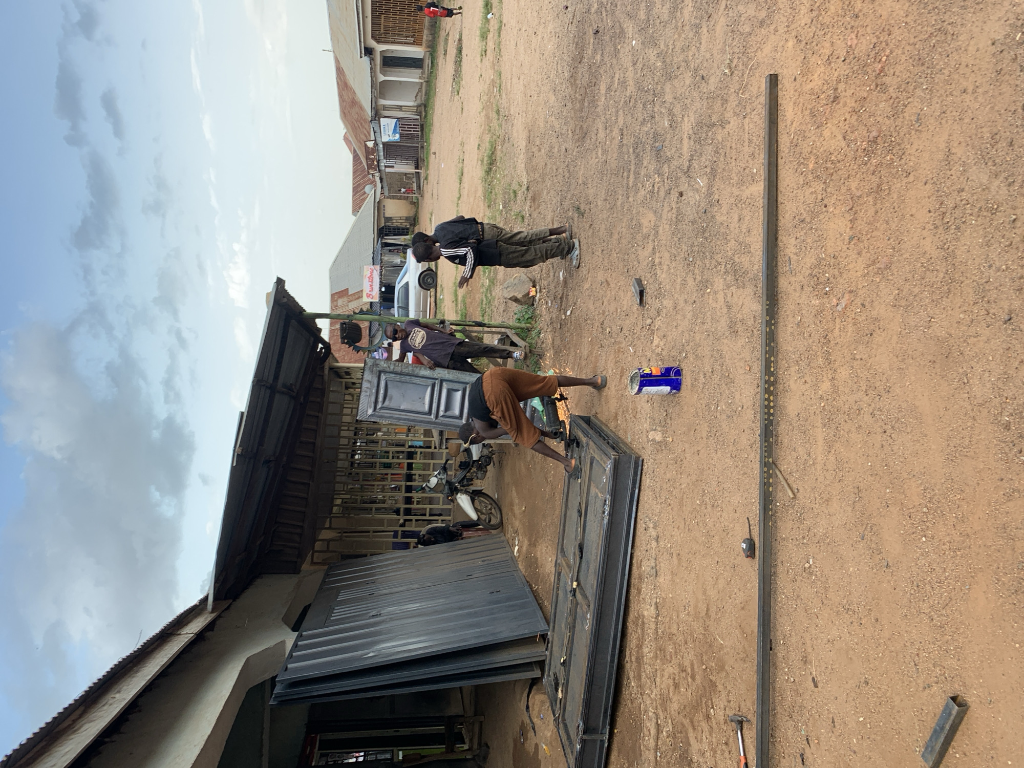 The image captures an outdoor scene in Jos, Nigeria, likely a workshop or marketplace area. The primary subjects are several men, seemingly engaged in metal fabrication. One man, wearing a black shirt and brown shorts, is actively working on a metal door frame, appearing to be welding or grinding, as indicated by sparks near his work. Another man in a purple shirt and sunglasses is observing. Further in the background, two men in casual attire stand near a white vehicle and some buildings.
The setting is characterized by a dusty, uneven ground with scattered debris. To the left, a rudimentary workshop structure with a corrugated metal roof is visible, displaying stacks of metal doors and frames. In the background, a row of buildings with awnings and some signage suggests a commercial area. A white car is parked in the middle distance, and a motorcycle is parked near the workshop.
The lighting and sky suggest it is daytime, possibly late morning or early afternoon, with partly cloudy skies. The overall atmosphere appears to be one of work and commerce.
Notable details include a blue and white can of paint or solvent lying on the ground near the working man, a metal pipe, a hammer, and a measuring tape scattered on the ground, all typical tools and materials in such a setting. There are no clearly legible signs with text that can be deciphered from the image.