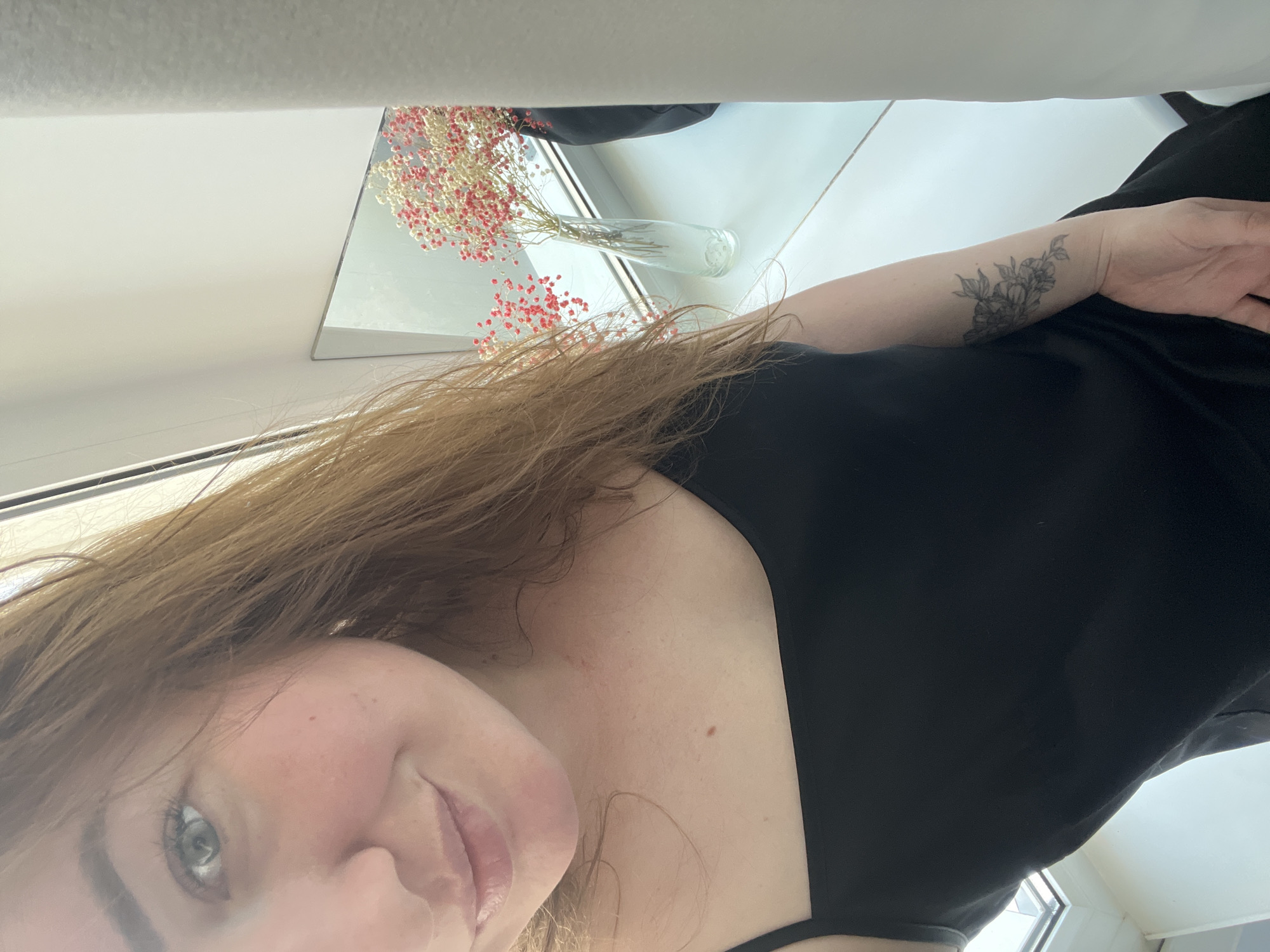 A female subject is captured in a selfie taken indoors in the city of Chaikovsky, Russia. The subject, possessing long, light brown hair, is positioned in the foreground, facing the camera with a gentle smile. Her left eye, green-blue in hue, is clearly visible. She is attired in a black sleeveless top with thin straps. A floral tattoo, rendered in dark ink and depicting flowers and foliage, is present on her left forearm. The background features a brightly lit white windowsill or shelf. On this surface, a slender, clear glass vase holds a dried floral arrangement characterized by numerous small, clustered blossoms, predominantly white with significant bright red portions. Behind the vase, a rectangular mirror leans against a light-colored wall, reflecting the flowers. A vertical white fabric element, likely a curtain or blind, partially obscures the right side of the scene. A window with a white frame is visible to the left, providing natural illumination.
