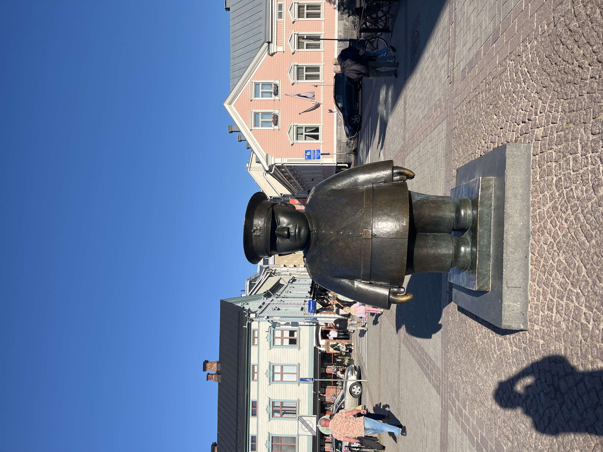 In the city of Oulu, Finland, a bronze statue of a uniformed figure stands prominently in a public square. The statue, depicting a person with a round face and wearing a cap and uniform, is positioned on a dark base. The square is paved with cobblestones and interspersed with patterned brickwork.
Surrounding the square are buildings with traditional architecture. To the left, a light-colored building with dark shutters and a dark roof is visible. Further down the street, other buildings with colorful facades, including a pink one with white trim, line the square. The sky above is a clear, bright blue.
Several people are present in the scene. On the left side of the square, a person in a patterned shirt and blue jeans walks away from the camera. In the background, near the buildings, other individuals are seen walking and interacting. Some cars are parked or driving in the background. The overall impression is a sunny day in a European city square.