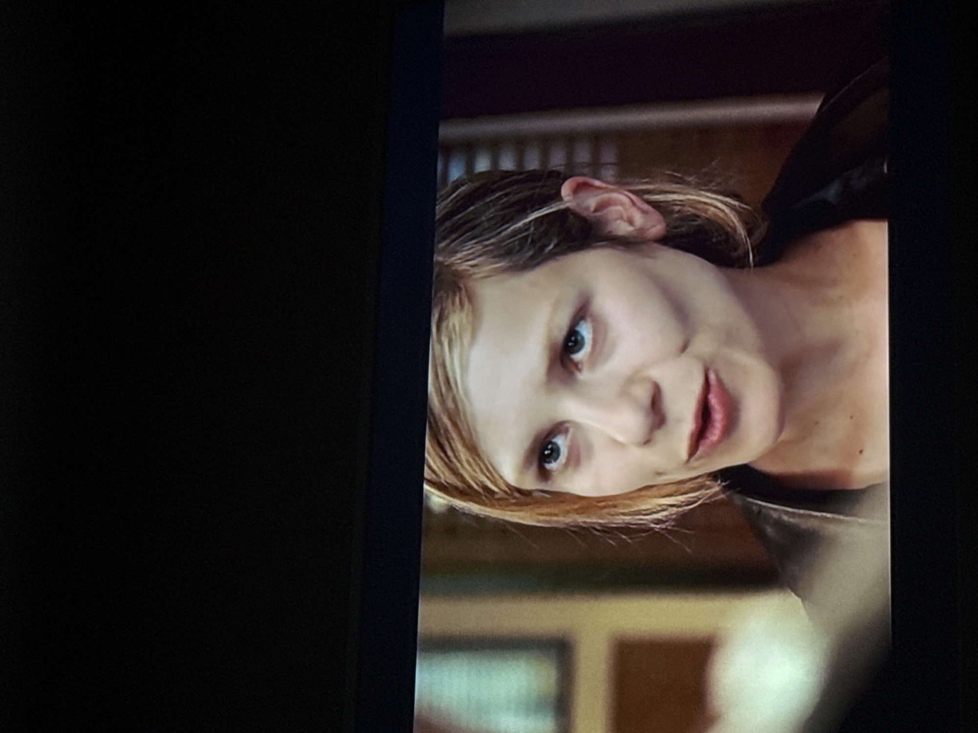 A medium close-up, slightly angled, shows a light-skinned woman with blonde-brown hair, facing the viewer. Her eyes are green-blue, and her lips are slightly parted. She is wearing a dark top. The background is blurred but shows what appears to be a room interior with wooden blinds. The image is displayed on a screen, with a black border on the left and right. The location in Mytishchi, Russia, is not evident in the image.