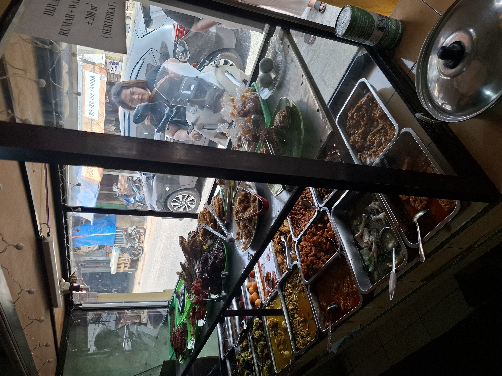 This image depicts a food stall in Cibadak, Indonesia, seen through a glass display case. The stall features an array of Indonesian dishes laid out on tiered shelves. These include various types of fried fish, curried dishes, grilled meats, and what appear to be seasoned vegetables. Several metal serving spoons are placed within the food trays.
In the background, visible through the glass, is a street scene with motorbikes and cars. A woman is seated on a white scooter, facing towards the food stall, suggesting she may be a customer or the stall owner. She is wearing a black t-shirt. A sign in the background reads "WR IBU IDA FARIDA," likely identifying the business. The environment appears to be outdoors, with a sunlit street. A can with text is visible to the right of the display.