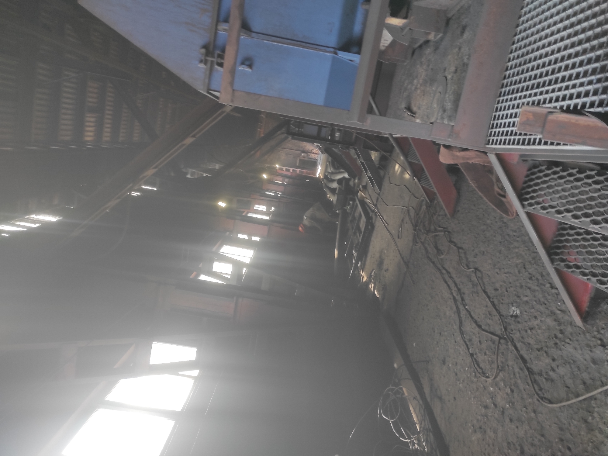 The image depicts the interior of an industrial facility, likely a workshop or factory, with a raw, unfinished aesthetic. Natural light streams in from large windows on the left, creating bright areas and casting long shadows. The floor is covered in a coarse, dusty material, possibly concrete or dirt, with various cables and wires strewn about. In the mid-ground, a person wearing an orange hard hat is bent over, seemingly engaged in some form of work or repair. To the right, a large blue metal structure, possibly part of machinery or equipment, dominates the scene. The background reveals a high ceiling with exposed beams and corrugated roofing. The overall atmosphere is one of active industrial work, with elements of construction or maintenance visible. The location is identified as the city of Kyzyl, Kazakhstan.