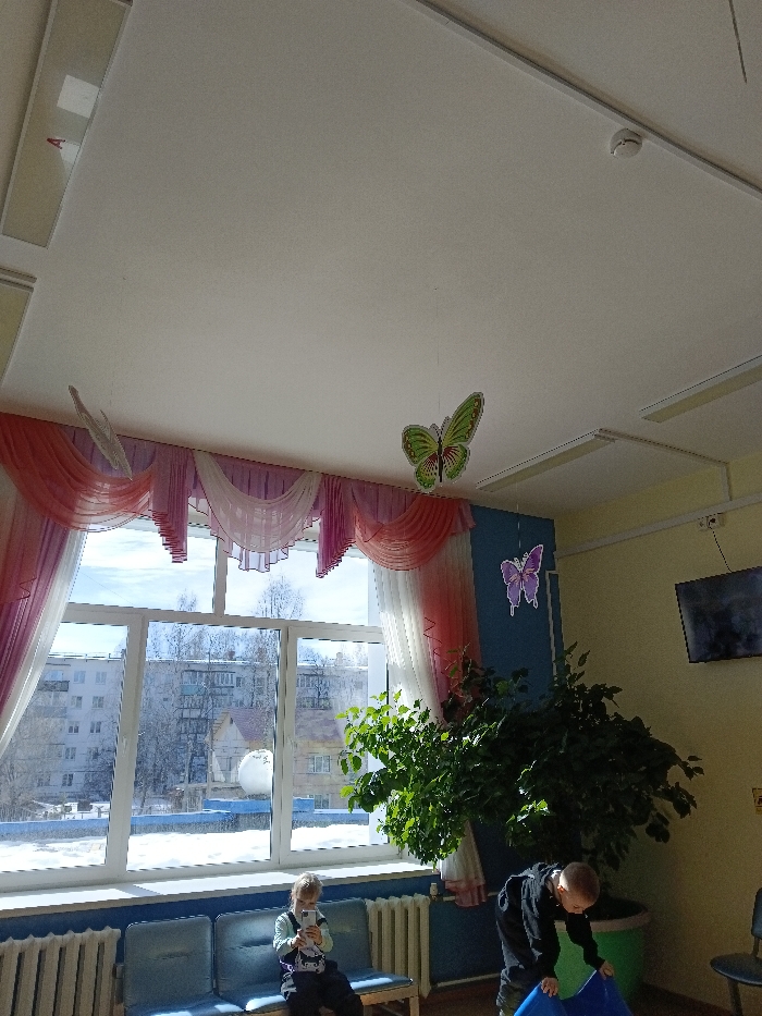 The image depicts an indoor scene, likely a waiting area or common room, in Chaikovsky, Russia, illuminated by natural light from a large window and overhead fluorescent panels.
**Environment:**
The room features a white ceiling with multiple rectangular fluorescent light fixtures and a circular smoke detector visible in the upper right. Three decorative butterflies are suspended from the ceiling: a large green one centrally, and a smaller, paler one to the left. A large, multi-paned white window dominates the far wall, dressed with layered sheer curtains in shades of pink, white, and purple. White horizontal panel radiators are installed beneath the window. To the right of the window, a section of the wall is painted blue, transitioning to a light yellow wall further right. A large, dark green potted plant stands to the right of the window, partially obscuring the curtain. A flat-screen television or monitor is mounted high on the yellow wall to the far right, displaying a dark image. The floor is light-colored in the foreground, darkening towards the right.
**Outdoor View (through the window):**
The view outside shows an urban landscape characteristic of Chaikovsky, Russia. Multi-story residential buildings, primarily light-colored, are visible in the mid-ground and distance. Bare deciduous trees with visible branches are scattered between the buildings, indicating a winter or early spring season. The ground appears partially covered with snow. The sky is bright with some cloud cover.
**People and Objects:**
1. **Child 1 (left):** A young girl, approximately 5-8 years old, with light-colored hair, is seated on a light blue upholstered bench in front of the window. She wears a light-colored long-sleeved shirt and dark pants with a white pattern on the leg. She holds a light blue smartphone in both hands, attentively focusing on its screen.
2. **Child 2 (right):** A young boy, approximately 6-9 years old, wearing a dark hooded sweatshirt and dark pants, stands near the large potted plant. He is bent over, interacting with a bright blue plastic object on the floor, which resembles a small play slide or ramp. His attention is directed downwards towards the object.
3. **Additional Decorations:** A smaller purple butterfly decoration is affixed to the blue-painted wall section to the right of the window.