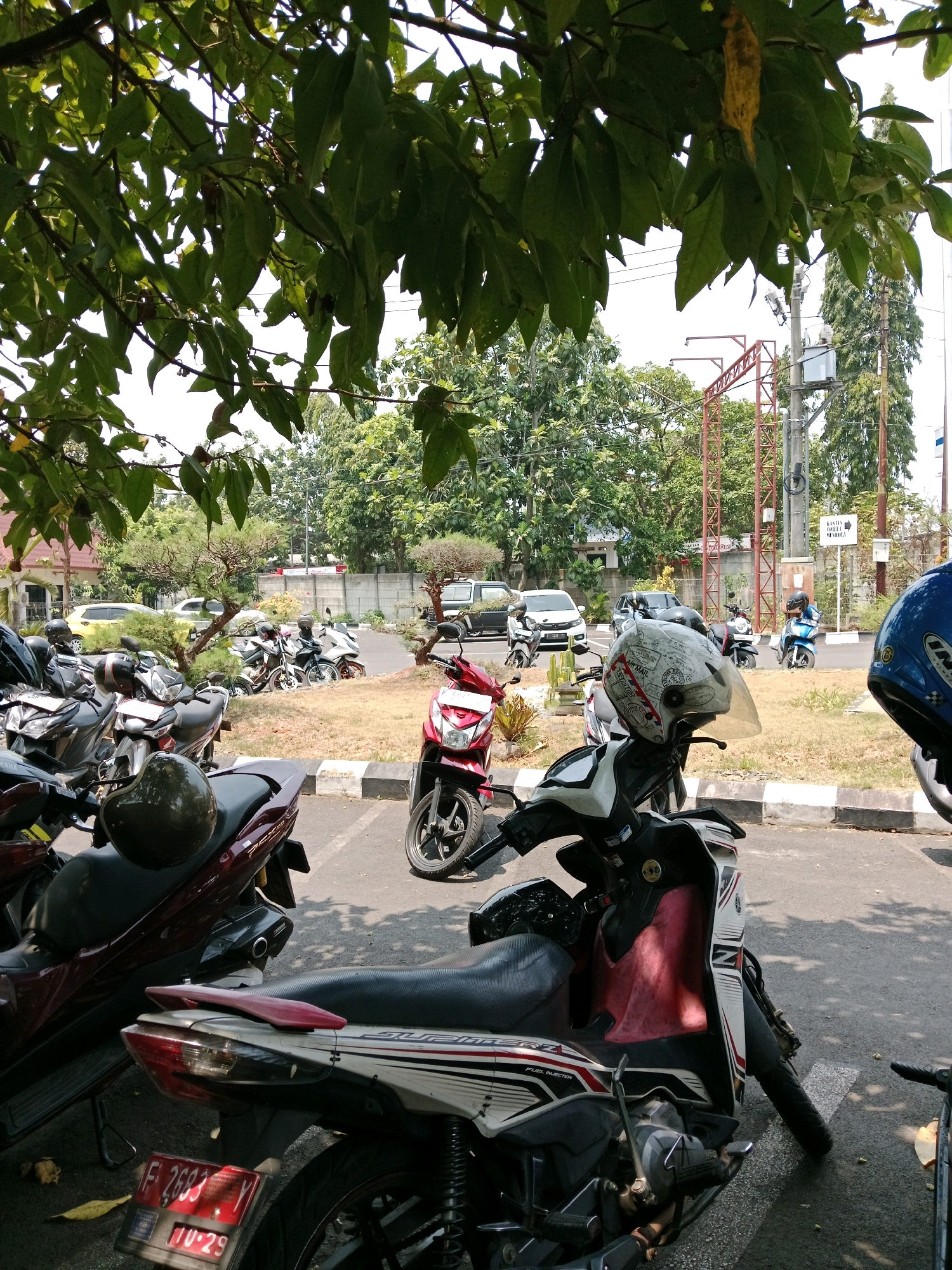 In the city of Karangtengah, Indonesia, a parking lot scene unfolds under the shade of lush green trees. The foreground is dominated by several parked motorcycles, their metallic bodies reflecting the sunlight. One maroon scooter in the lower left has a dark green helmet resting on its seat. Adjacent to it, a white and red scooter is parked, its front wheel turned slightly. Further into the mid-ground, a red scooter with a rider in a blue shirt is visible, parked beside other motorcycles. In the background, a white car and a yellow car are parked on a paved road. A concrete barrier with grass and small trees separates the parking area from the road. In the far background, a metal structure, possibly part of a construction or utility site, stands against a backdrop of dense green trees. Electrical poles and wires are also present in the scene. The overall environment appears to be a public space, likely a parking area for a commercial or administrative building.
