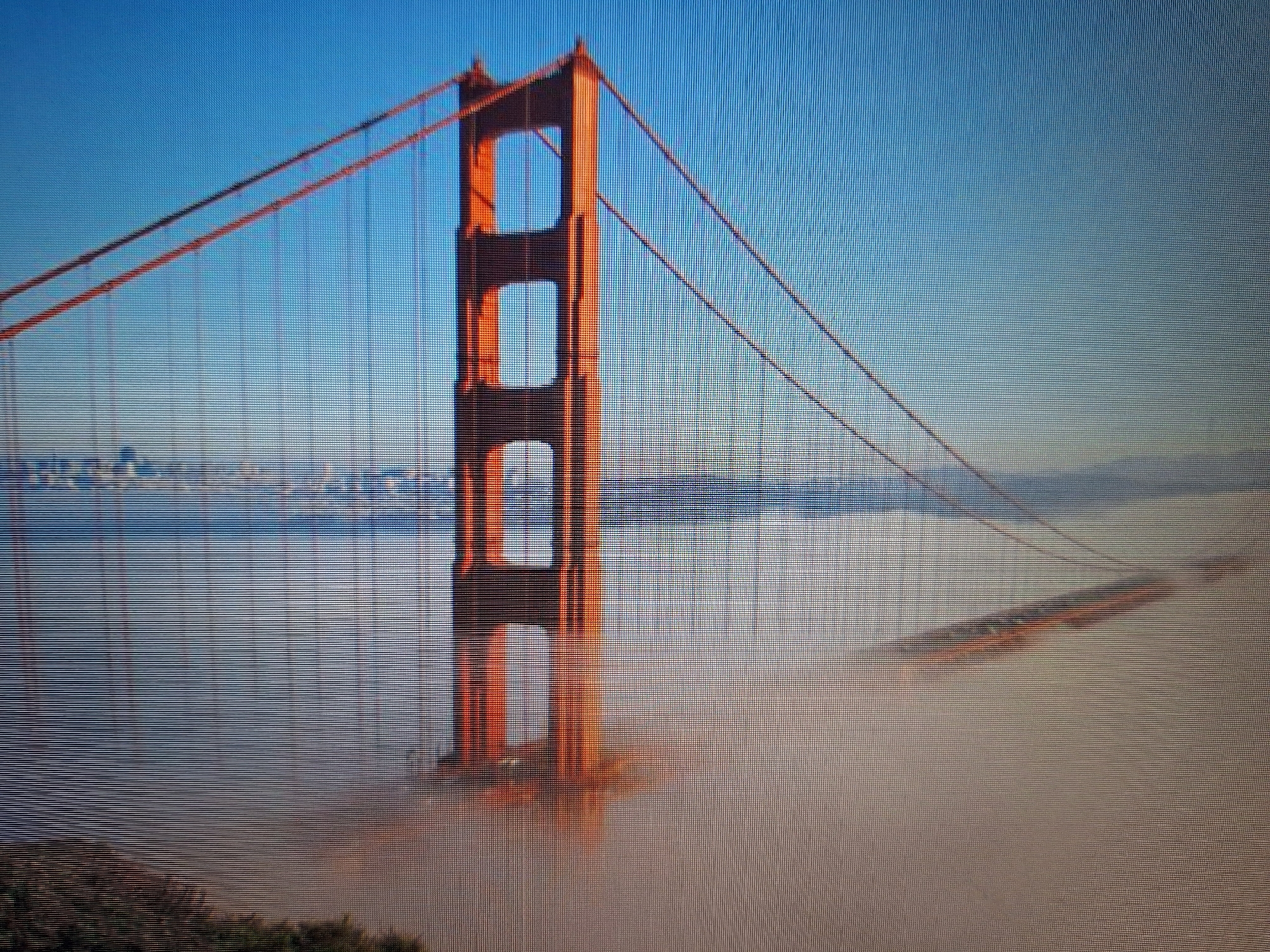 The image shows a wide-angle view of the Golden Gate Bridge, shrouded in fog. The iconic orange towers of the bridge are visible, along with its suspension cables. The fog obscures the lower portions of the bridge and the water below, creating a dramatic and atmospheric scene. The sky above is clear and blue. In the distance, the faint outline of a city skyline can be seen through the fog. The image appears to be taken from a slightly elevated position, looking towards the bridge. There are no people or animals visible in the image. The location is not Varna, Bulgaria, but rather San Francisco, California.