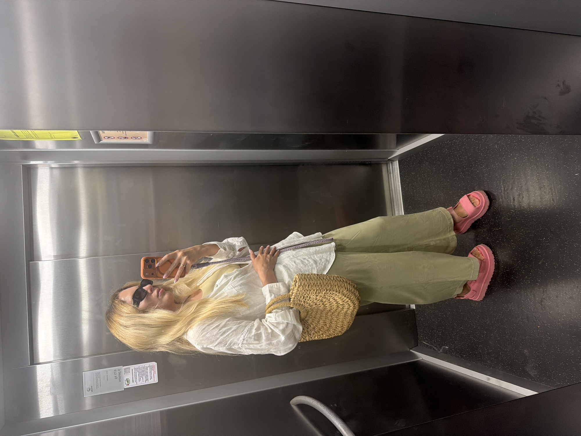 A woman with blonde hair and sunglasses is taking a mirror selfie in an elevator. She is wearing a white linen shirt, a yellow camisole, olive green wide-leg pants, and bright pink platform sandals. She is carrying a woven straw bag on her left shoulder. The elevator has stainless steel walls and a black speckled floor. There are signs on the elevator walls, including one with a diagram of a person. The location is an elevator in Budva, Montenegro.