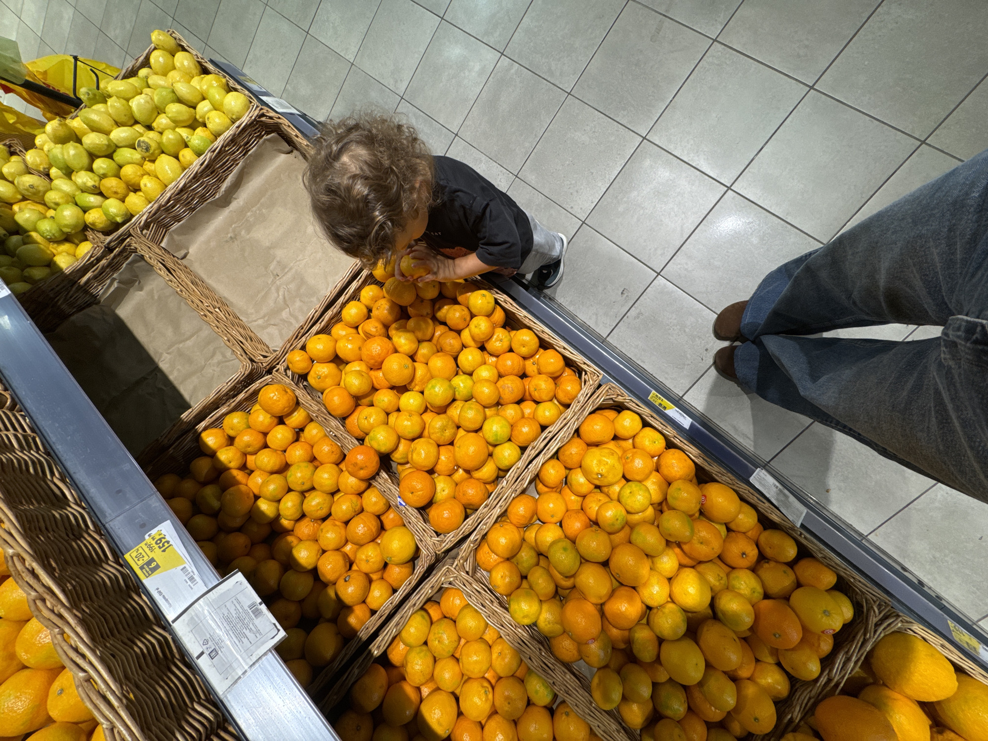 In a grocery store in Sochi, Russia, a young child with curly brown hair is reaching into a basket of mandarins. The child is wearing a dark long-sleeved shirt and grey pants, with dark shoes. The child is holding a mandarin to their face, possibly smelling or tasting it. The basket is part of a display of fruits, with other baskets filled with mandarins and lemons visible. The flooring is light gray tile. In the foreground, the legs of an adult wearing blue jeans and brown shoes are visible, indicating the perspective of the photographer. A price tag is visible on the edge of one of the fruit baskets.