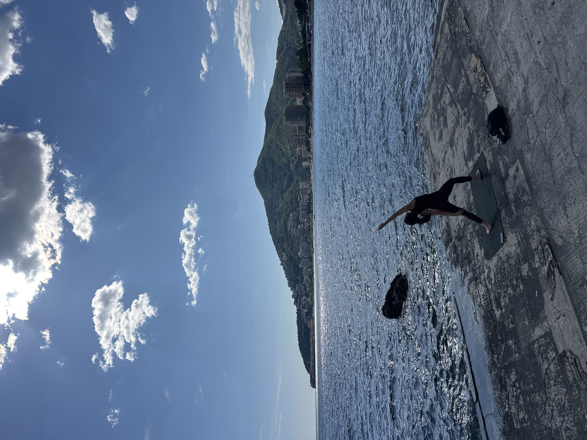 The image, when correctly oriented, depicts an outdoor scene by the sea, characteristic of Budva, Montenegro. A silhouetted figure, dressed in dark athletic wear, is performing *Trikonasana* (Triangle Pose) on a green yoga mat positioned on a concrete pier or platform. Her arms are extended, one reaching upwards and the other downwards, in a posture of balance and focus. A dark object, possibly a bag, rests on the concrete near the mat, and a rough, dark rock is partially submerged in the water close to the platform's edge.
Beyond the sparkling blue water, a large mountain covered in lush greenery and buildings rises in the background. The sky is a clear, vibrant blue with scattered white cumulus clouds, indicating bright and sunny weather. Strong sunlight creates dazzling reflections across the water's surface. The clear conditions and serene activity suggest a peaceful morning or late afternoon. There is no visible text in the media.
