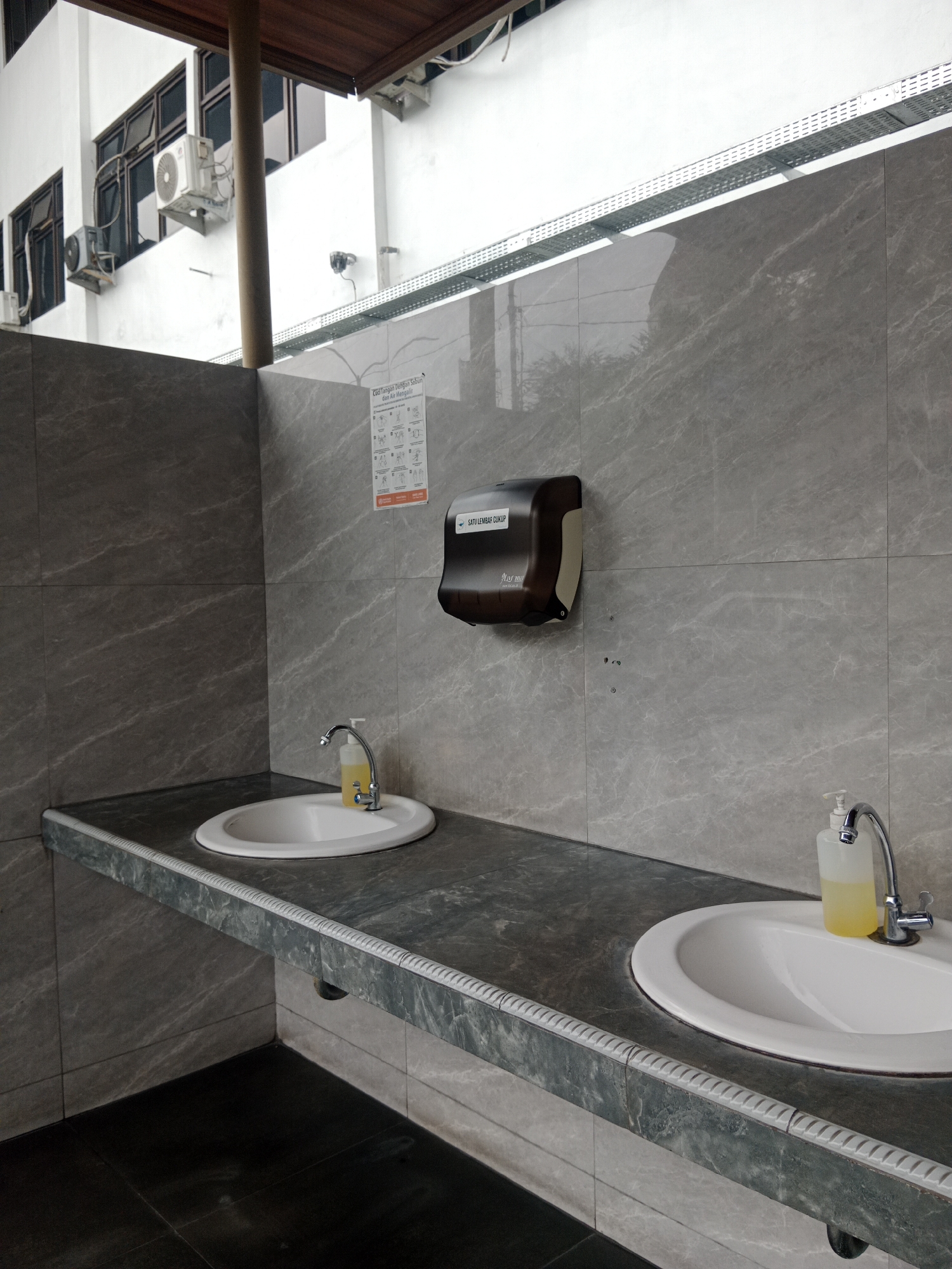 The image shows a public restroom area with two sinks. The walls are tiled with a gray, marble-like pattern. Above the sinks, there is a dispenser that likely holds paper towels or soap. There is a sign on the wall showing instructions for washing hands. The floor is tiled with dark squares. In the background, the exterior of a building is visible, with windows and air conditioning units. The overall impression is of a clean, functional public restroom. This scene is likely located in Yogyakarta, Indonesia, given the Indonesian text on the handwashing instructions sign.