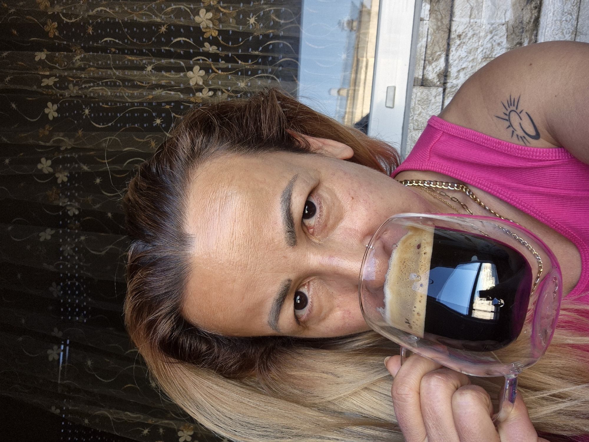 A medium shot captures a woman with blonde and brown hair, looking directly at the camera while holding a clear glass mug of dark coffee with a frothy top. She is wearing a bright pink tank top and a gold chain necklace. A sun tattoo is visible on her right arm. The background consists of a dark patterned curtain and a window with a reflection of buildings and sky. The scene appears to be set in an urban environment, possibly on a balcony or near a window, in Unknown, Unknown.