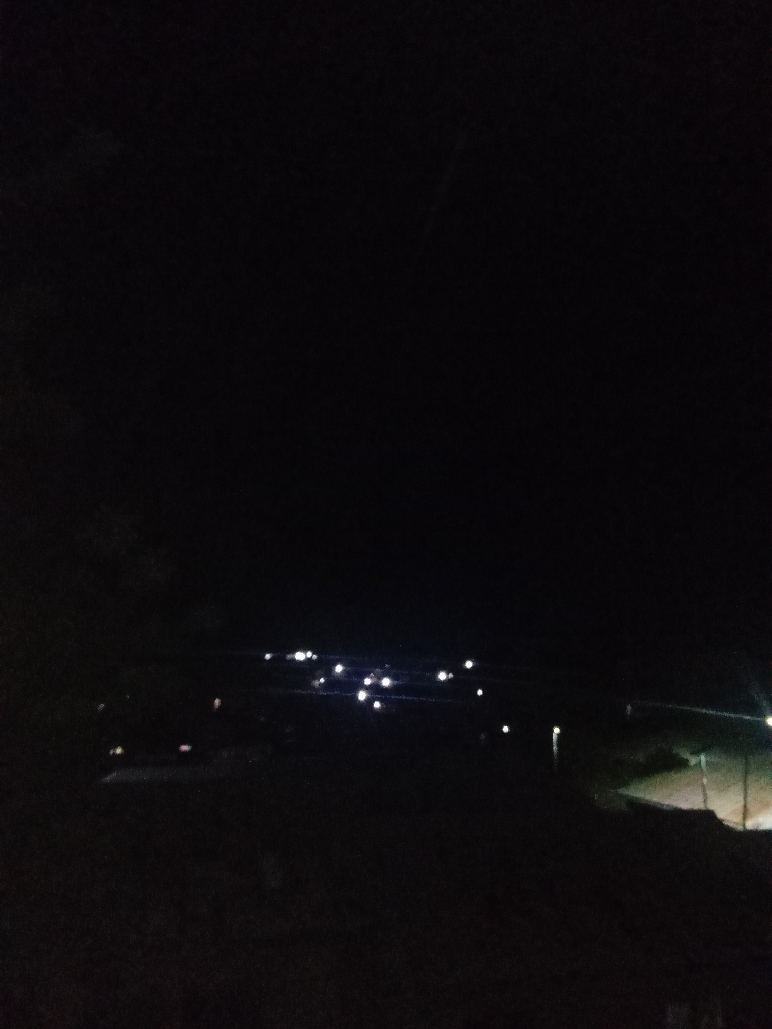 The image depicts a nighttime scene with a dark sky. In the lower portion of the frame, a series of lights are visible, suggesting the presence of a settlement or a road with streetlights. The lights appear scattered and some are elongated, indicating they could be from buildings, street lamps, or vehicles. The overall environment appears to be outdoors, with a dark, possibly natural landscape or unlit areas interspersed with the illuminated points. Without specific landmarks or recognizable structures, it is not possible to definitively identify the location within Lokoja, Nigeria.