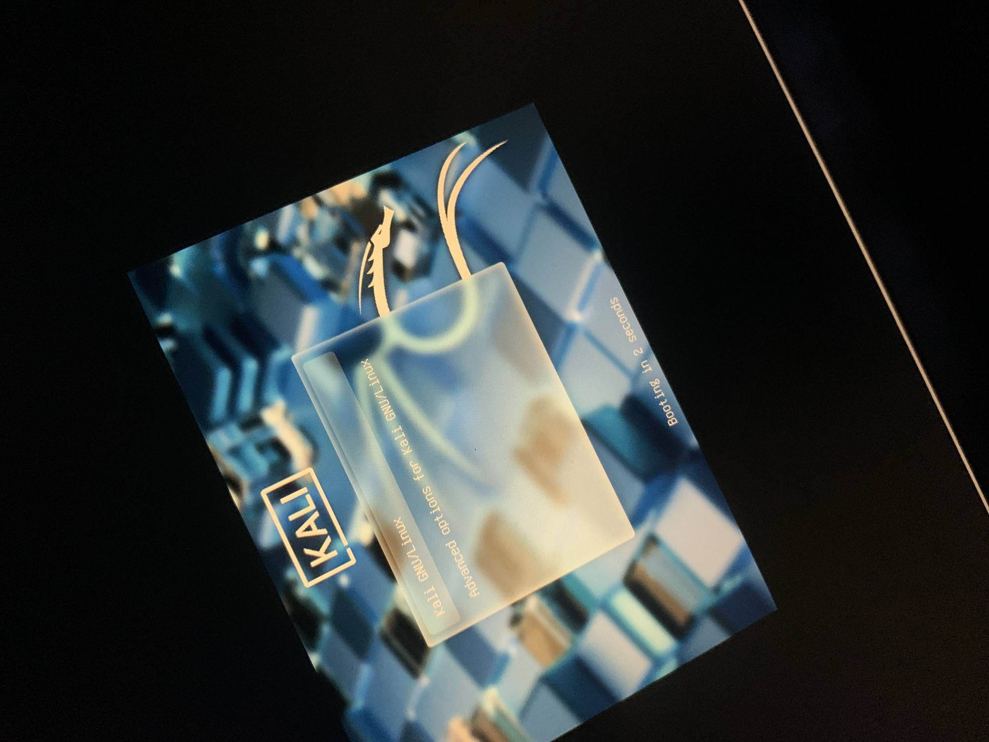 The image shows a screen displaying the Kali Linux boot menu. The Kali Linux logo, a stylized dragon, is visible. The boot menu options "Kali GNU/Linux" and "Advanced options for Kali GNU/Linux" are present. A message indicates "Booting in 2 seconds." The background of the screen is a blurred blue and white geometric pattern. The scene appears to be a close-up shot of a computer screen displaying the Kali Linux operating system booting up.