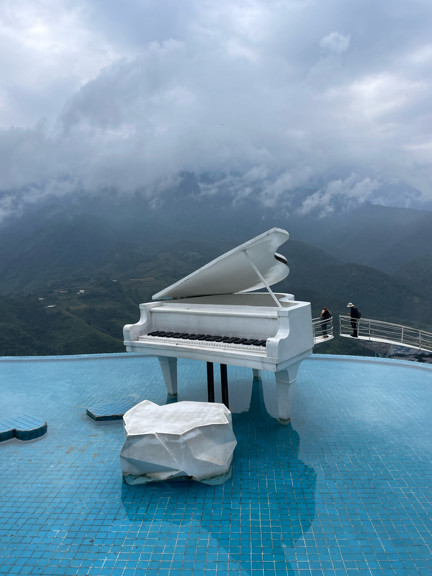 The image depicts a grand piano situated in a shallow, tiled pool with a cloudy, mountainous landscape in the background. The piano is white and appears to be a replica or artistic installation, as it is partially submerged in the blue water. A large, white, geometric rock sculpture is placed in the foreground, also within the pool. In the middle ground, two individuals are visible standing on a white metal walkway, observing the scenery. The environment is characterized by lush green mountains shrouded in mist and clouds, suggesting a humid or overcast day. The tiled pool extends towards the viewer, reflecting the sky and the piano. The scene appears to be a tourist attraction or viewpoint, likely in an elevated location offering panoramic views of the Sa Pa region in Vietnam.