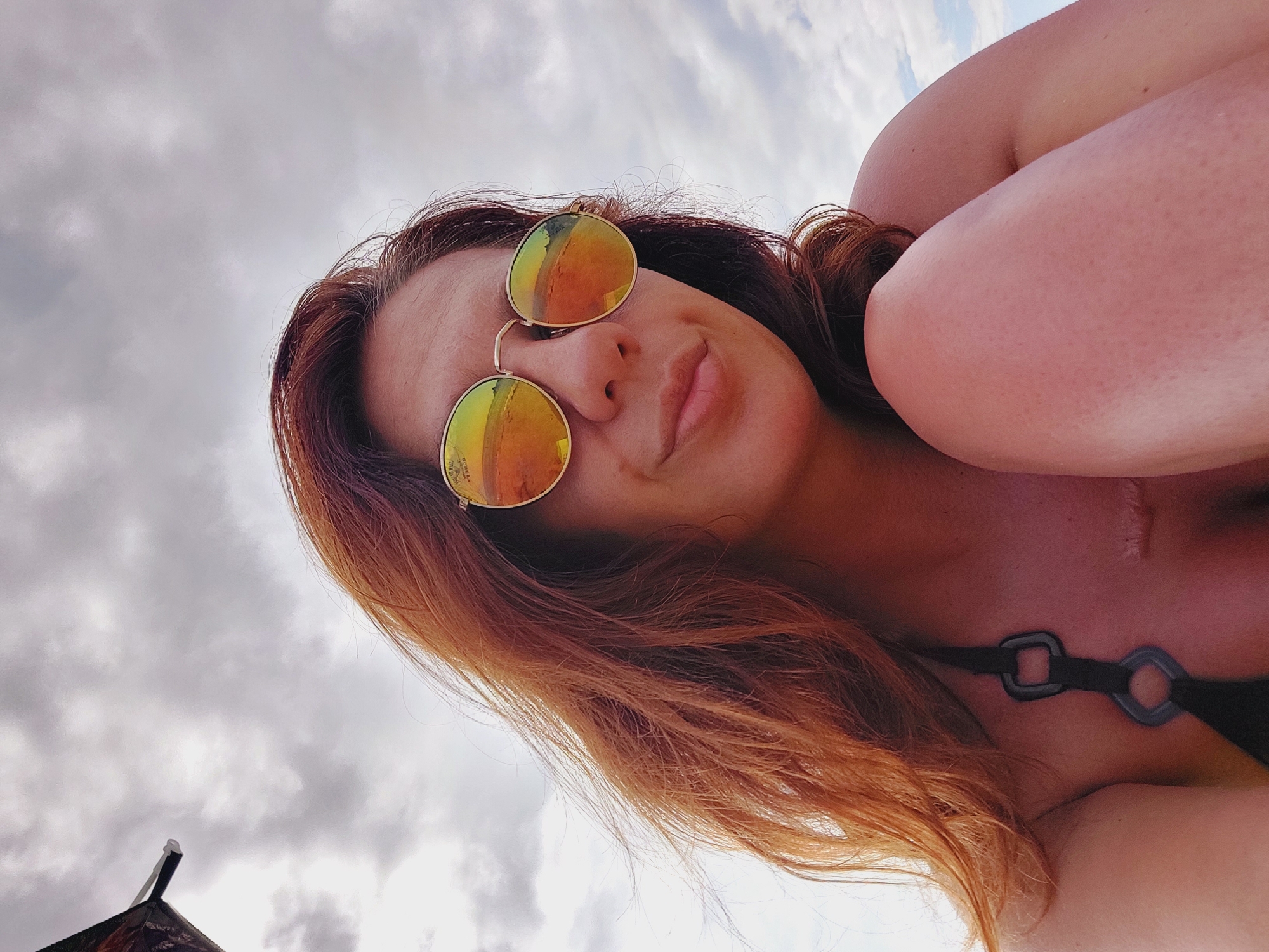 A low-angle, close-up, outdoor selfie of a woman with auburn hair and sunglasses is taken against a cloudy sky. The woman is wearing a black bikini top. Her left shoulder and arm are visible, as well as part of her leg. A scar is visible on her chest. The background consists of a gray, overcast sky with some patches of lighter clouds. The woman appears to be at a beach, as suggested by the reflection in her sunglasses, which shows a sandy area and possibly the sea. The overall impression is a casual, summery portrait.