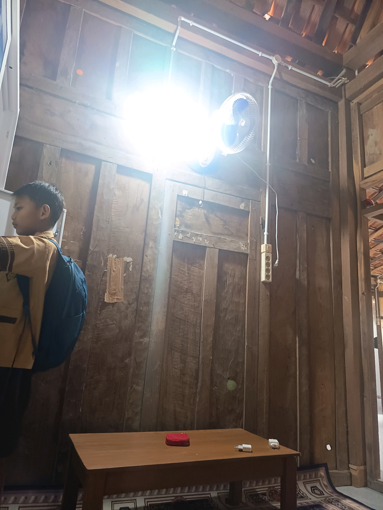 In Sidorejo, Indonesia, a boy wearing a tan uniform and a blue backpack stands in what appears to be a rustic wooden room. He is positioned on the left side of the frame, facing away from the viewer, looking towards a bright light source. The room's walls are constructed from wooden planks. Above the boy, a ceiling fan is mounted on the wall, and a bright light illuminates the scene, obscuring details in the upper portion of the image. Below the boy, a light brown wooden table is visible. On the table, a small red object, possibly a book or a case, and two white electrical adapters are present. The floor is covered by a patterned rug. Electrical conduits and a power strip with multiple outlets are attached to the wall. The overall impression is of an indoor space with a simple, traditional aesthetic, possibly a classroom or a common area within a residence.
