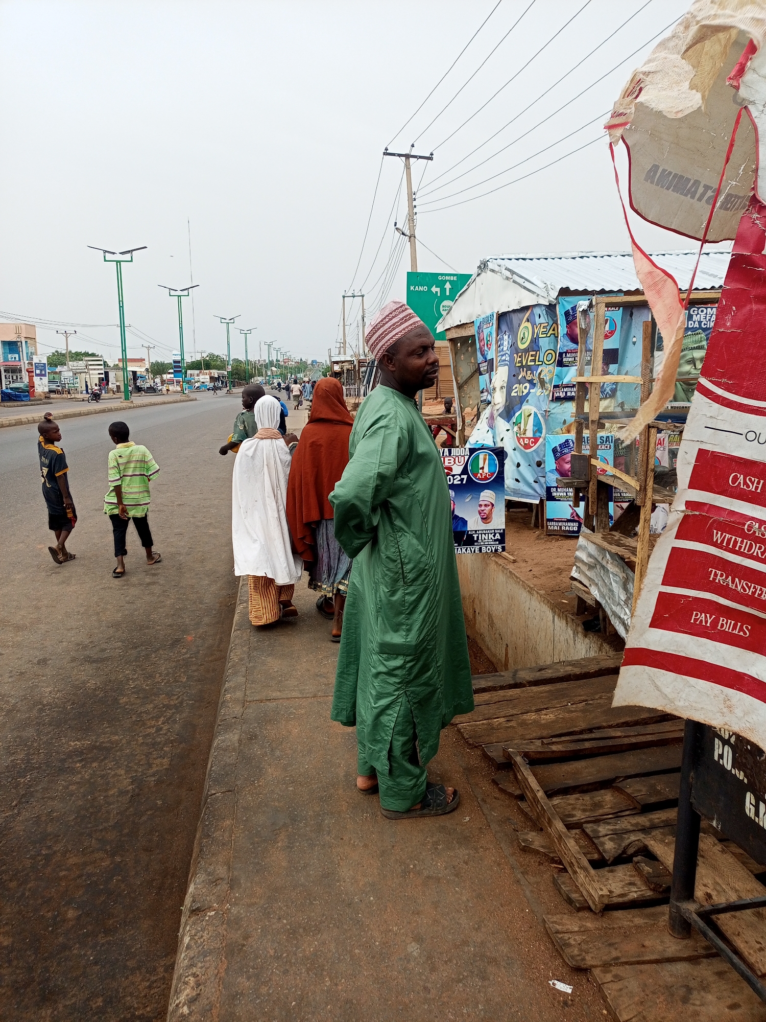 The image shows a street scene with several people, a roadside business, and political posters in an Unknown location. A man dressed in a green robe and cap stands near a roadside stall covered in political posters, which include images of candidates and the acronym "APC". A ripped and faded poster obscures part of the stall, displaying the words "CASH", "WITHDRAW", "TRANSFE", and "PAY BILLS". On the left of the frame, a few children are walking on the road. Behind them, other people, including one dressed in white and another in brown, are walking on the sidewalk. A green road sign pointing towards "GOMBE" and "KANO" is visible in the background. The scene is set on an overcast day, with utility poles and overhead wires stretching across the sky. Modern street lights line the road. The business stall has wooden planks on the ground.